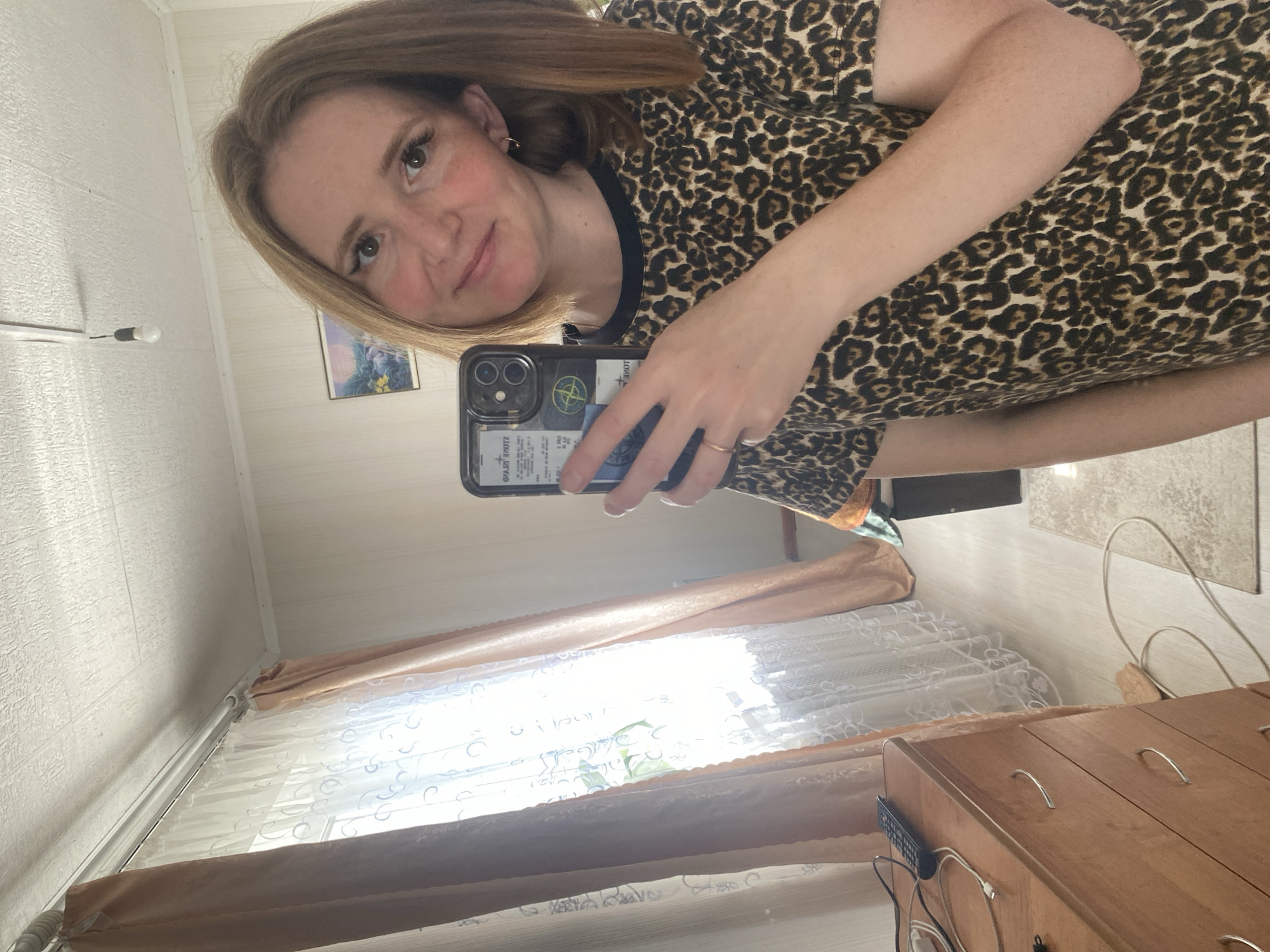 This is a vertical, slightly angled selfie of a young woman taken indoors. She has long, reddish-brown hair and fair skin with freckles. She is wearing a short-sleeved dress with a black leopard print and a black neckline. Her left arm is visible, resting on her side. She is holding a smartphone in her right hand, which is angled to capture the reflection in what appears to be a mirror, as the image is a mirror selfie. The phone case is black with some stickers, one of which appears to be a circular logo with text and graphics. A thin gold ring is visible on her ring finger.
The background shows an interior scene. To the left, a textured white wall is visible, with a simple lamp or fixture attached. Further back, a framed picture hangs on the wall, depicting a natural scene with flowers. A window with light-colored curtains, one sheer and one opaque, is prominent in the lower half of the image, suggesting it's daytime. Beyond the window, the sunlight is bright, indicating it's likely a sunny day. To the right, a wooden dresser or cabinet with silver handles is partially visible. The overall setting appears to be a domestic environment, possibly a bedroom or living area. The woman's expression is relaxed and pleasant. There is no visible text in the image itself that is clearly legible and relevant to the scene. The location context of Chaikovsky, Russia is provided externally.