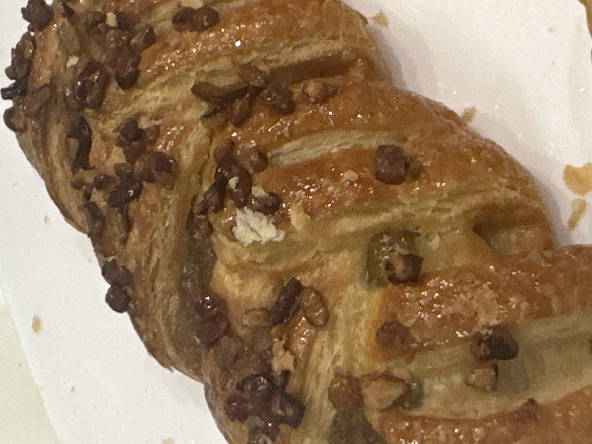 A close-up, high-angle forensic view captures a single, braided pastry resting on a white, undifferentiated surface. The pastry exhibits a golden-brown, glossy exterior, coated with a clear glaze and scattered with numerous irregularly shaped, dark brown chopped nuts and some shimmering sugar crystals. Its laminated structure is visible, showing distinct layers characteristic of a Danish pastry. Scattered crumbs are present on the white surface around the pastry. The scene is located within the city of London, United Kingdom.