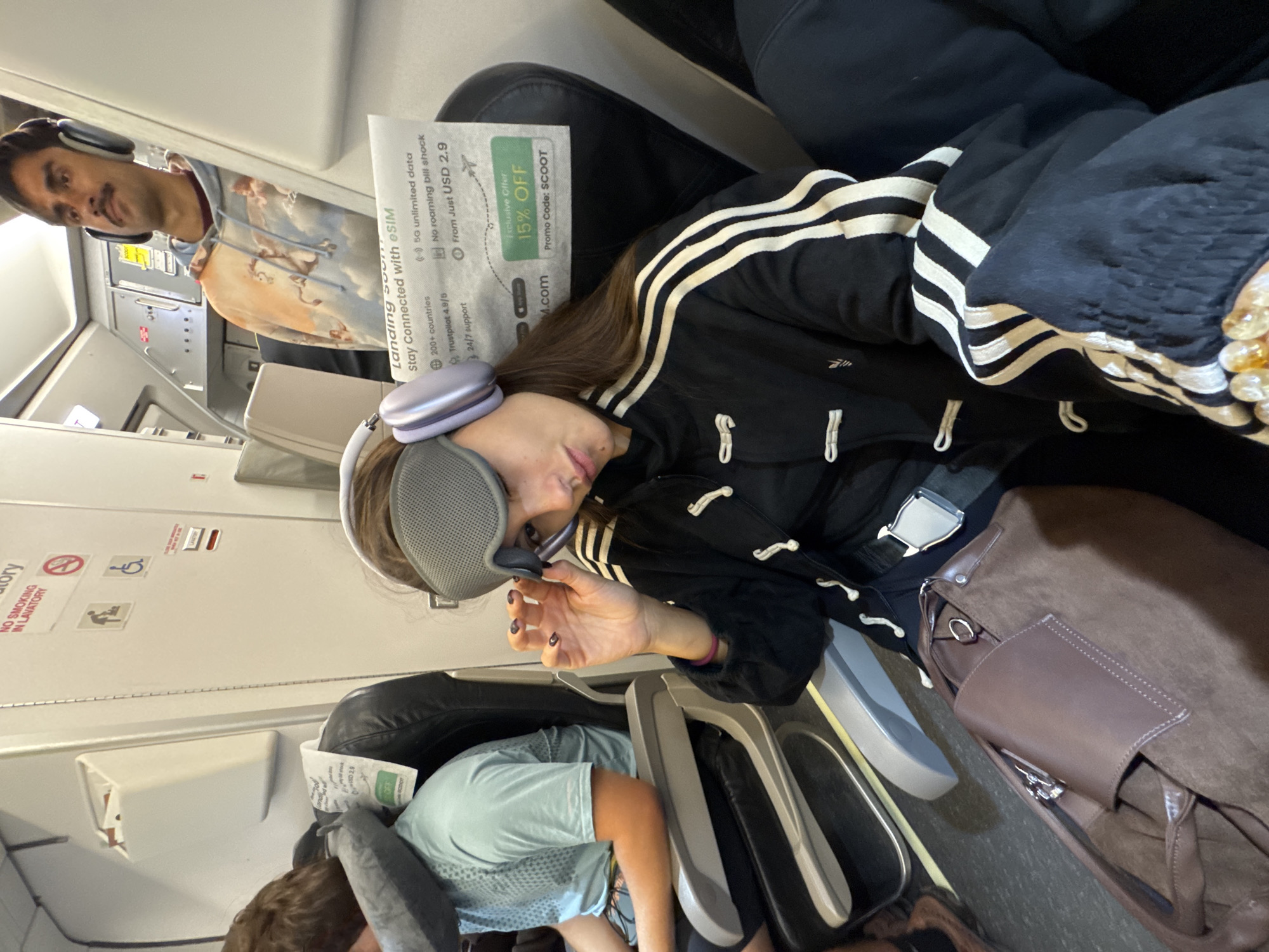 Here is a detailed description of the image:
The image shows an airplane cabin interior. A woman is seated in a window seat, wearing a black Adidas jacket with white stripes, an eye mask, and headphones. She appears to be trying to sleep. Next to her is a brown handbag.
In the next seat toward the aisle, a child wearing a light blue shirt is also trying to rest. A man in a hoodie and headphones can be seen across the aisle and slightly behind the woman.
The interior of the aircraft cabin is visible, including the overhead compartments, seats, and emergency exit signs. Promotional material for eSIM services and Scoot airlines is tucked into the seat in front of the woman. The aircraft appears to be landing soon, indicated by signage.
Based on the promotional material for Scoot airlines, the location could potentially be near the Phuket International Airport in ตำบล ไม้ขาว, Thailand, as Scoot is a major airline operating from this airport.