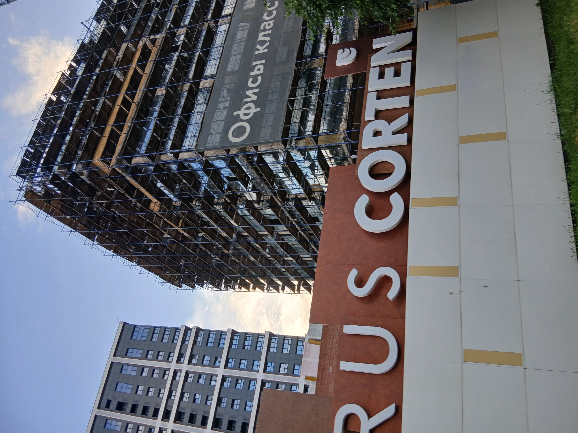 The image shows a construction site in Izhevsk, Russia, with two large buildings. On the left is a completed modern residential building with a dark facade and many windows. On the right is a building under construction, covered in scaffolding and a large banner displaying the Russian text "Офисы класс" (Offices class). In the foreground, there is a reddish-brown structure with large white letters spelling out "RUS CORTEN". Below this sign is a white wall with yellow vertical accents. The sky above is blue with some clouds. There are no people visible in the image.