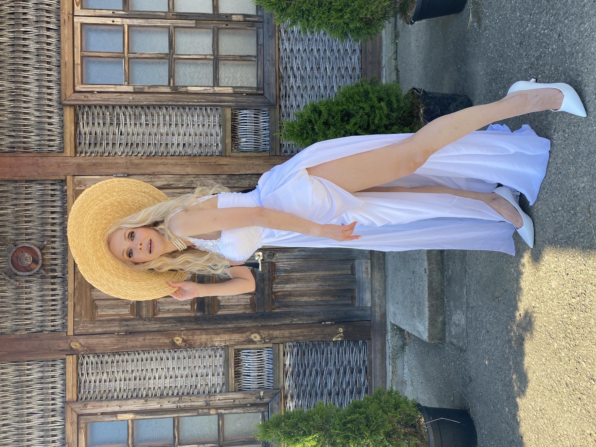 A fair-skinned woman with long blonde hair, wearing a wide-brimmed straw hat, a white sleeveless gown with a high slit, and white pointed-toe heels, poses in front of a rustic wooden building with wicker accents. She is standing with her left leg bent and extended, her right hand resting on her hat. Two evergreen bushes in black pots flank the building. The building's facade features wooden doors and windows with glass panes. The ground is paved with concrete. The image is an outdoor daytime shot, likely taken in Sochi, Russia, given the architectural style and vegetation.
