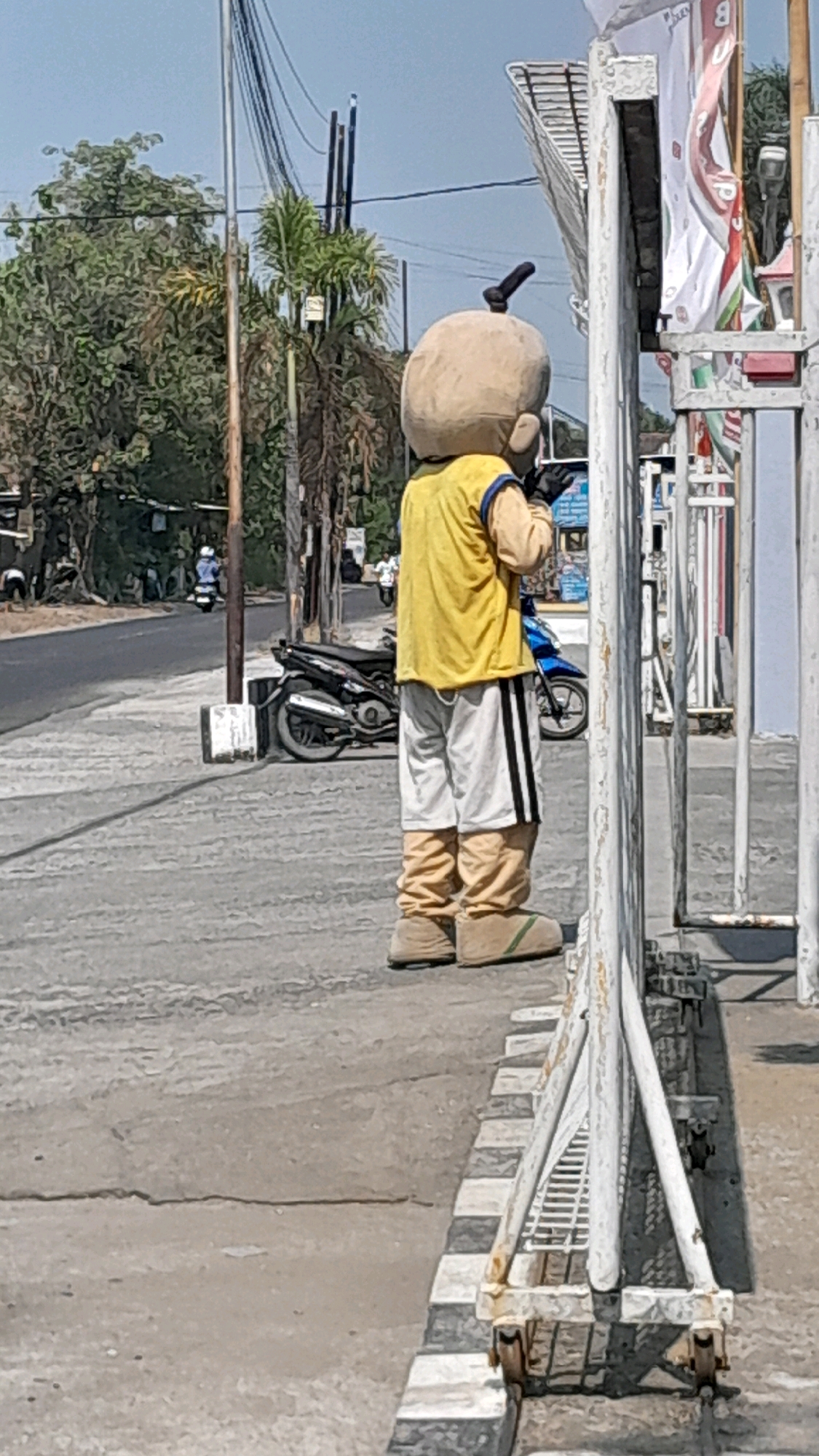 A person in a costume resembling a baby with a large head and a black knot on top stands on a sidewalk in Tanjung, Indonesia. The costume includes a yellow shirt with blue trim, white shorts with black stripes, and tan boot-like shoes. The person is wearing black gloves and appears to be holding a cell phone to their ear. Behind the person, a black motorcycle is parked on the side of a paved road. The environment is sunny with trees and utility poles visible in the background. A white metal fence with checkered black and white markings along the base separates the sidewalk from the street.