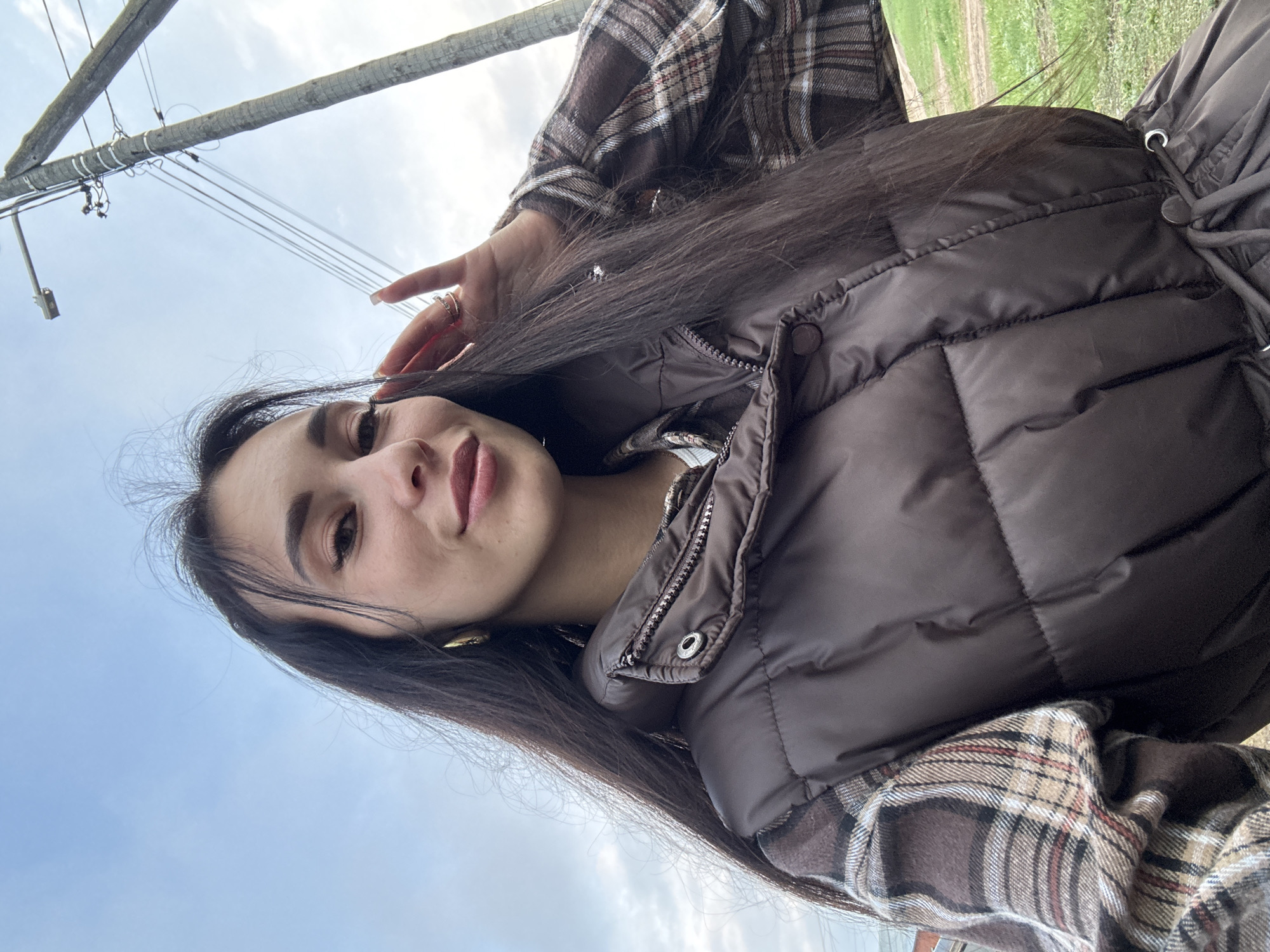 A young woman is taking a selfie outdoors, likely in Chaikovsky, Russia. She is wearing a brown puffer jacket over a brown plaid shirt. Her long, dark hair is loose. The background shows a blue sky, a utility pole with wires, and a patch of greenery, possibly a field or roadside vegetation. The camera angle is low, looking up at the woman.