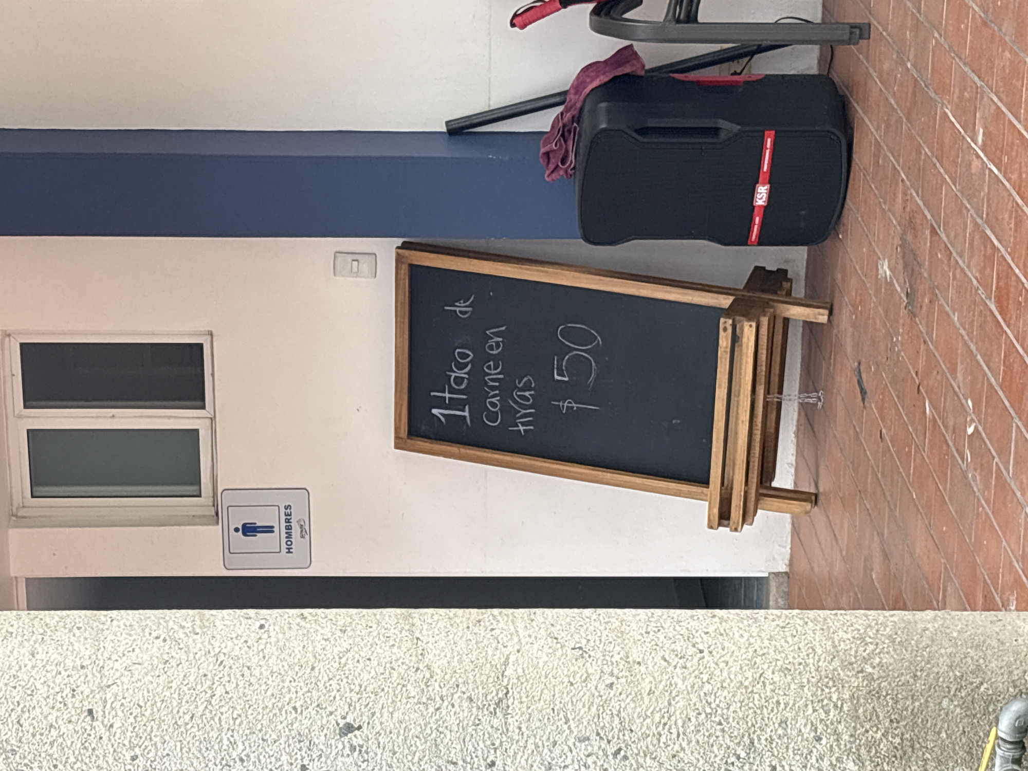 The image presents an outdoor scene adjacent to a light-colored building in San Francisco Totimehuacán, Mexico. A window is visible on the building's wall along with a men's restroom sign ("HOMBRES"). Adjacent to the restroom is a chalkboard sign, propped on a wooden easel, advertising "1 taco de carne en tiras $50" (1 taco of sliced meat $50). A black KSR speaker, partially covered with a dark cloth, sits on the ground and appears to be supporting the easel. The ground is divided between textured concrete paving in the foreground and brick paving in the background.