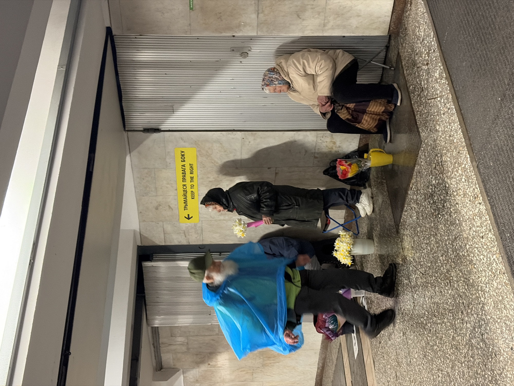 Here is a forensic description of the image:
The image depicts three individuals inside what appears to be an underground pedestrian area, potentially a subway station. The location is in Unknown, Unknown.
Nearest the camera, an older male is dressed in a blue poncho, dark trousers, and dark shoes. He has a white beard and wears a cap. A bucket of flowers is located near his feet, which he is either selling or carrying.
In the middle of the frame, a person in a black jacket and dark trousers is holding a small bouquet of flowers, with another bucket of flowers near their feet.
Further away, an older female is seated, wearing a beige jacket and headscarf. Next to her is a bucket with flowers.
A yellow directional sign is visible, written in a non-English script and English, stating "KEEP TO THE RIGHT". The background consists of metallic or tiled walls. The floor is speckled.
The individuals are either selling flowers or potentially seeking shelter in the underground space.