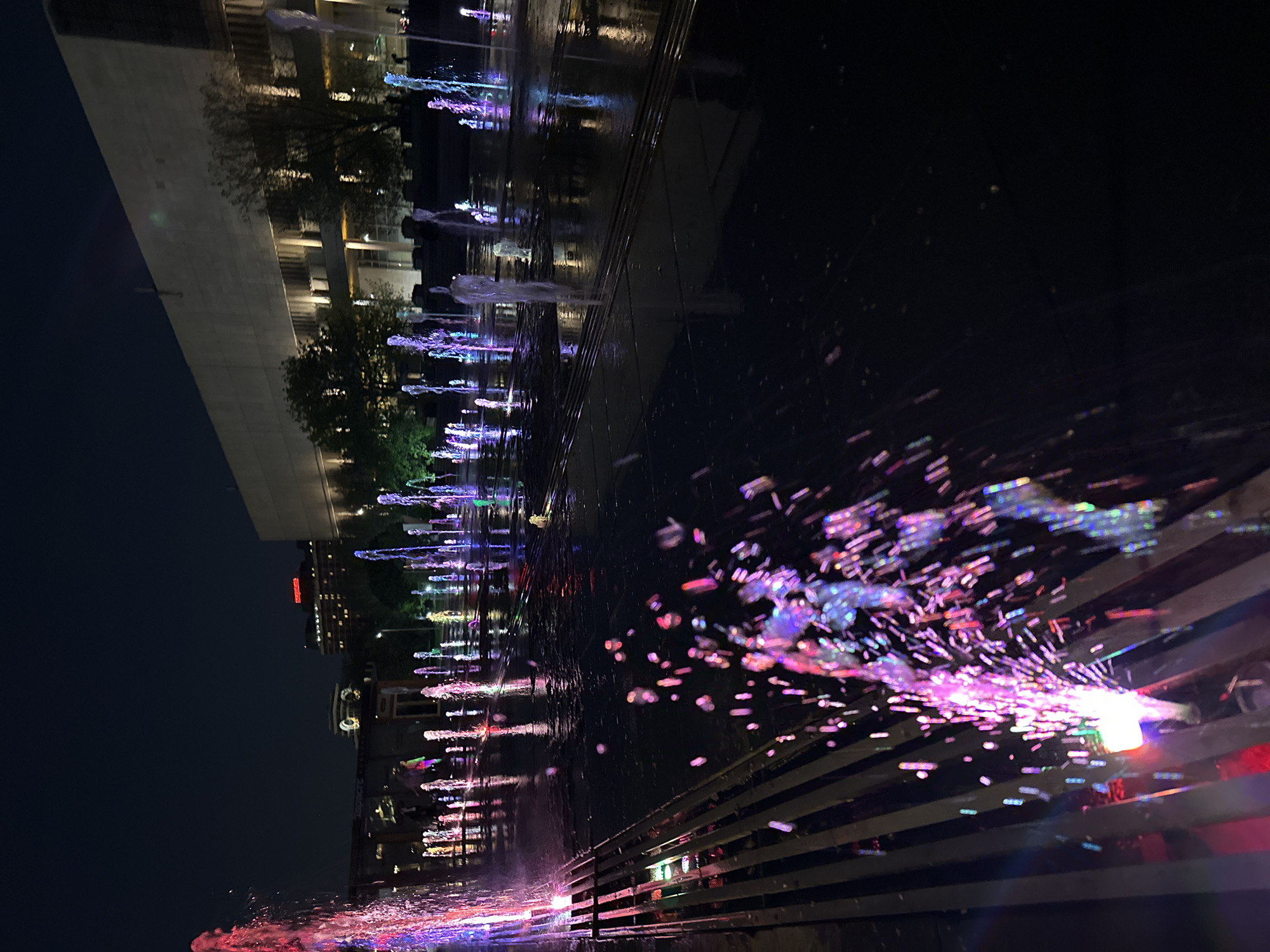 The image shows a water fountain at night, likely in an urban area. The fountain is illuminated with colorful lights, predominantly red, purple, and blue. Water jets shoot upwards and outwards, creating a dynamic display. In the background, buildings and trees are visible, reflecting in the water. The image is taken from a low angle, emphasizing the height and spread of the fountain. The overall scene is vibrant and captures the beauty of an illuminated fountain at night. Location is Unknown, Unknown.