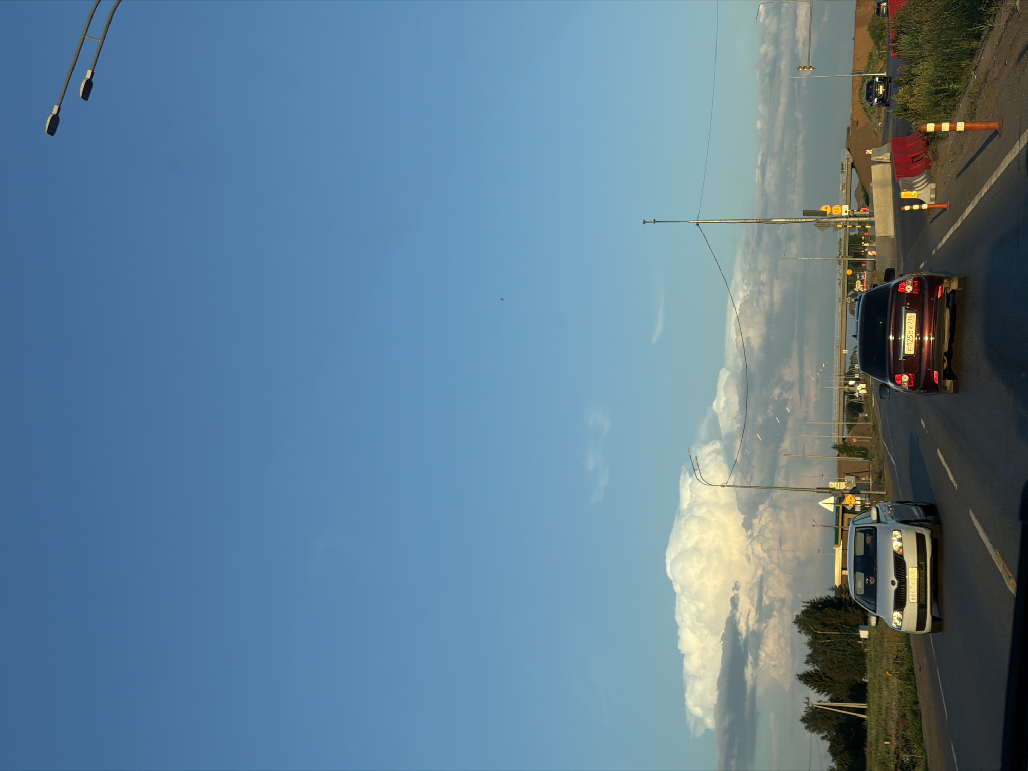 This image depicts a view from a vehicle traveling on a highway in Derzhavino, Russia. The sky is a clear, bright blue with a few wispy clouds scattered across the upper portion, and a large, towering cumulus cloud formation is visible on the horizon. The road is paved and has lane markings. In the foreground, two cars are on the highway. The car closest to the viewer is a dark red sedan with license plate E125OK716. Ahead of it is a white sedan with a license plate that appears to read B667HH49RUS. The road is lined with grassy verges and trees. In the distance, there are streetlights, overpasses, and construction barriers, indicating ongoing roadwork or infrastructure development. The overall scene suggests a summer day with good visibility.