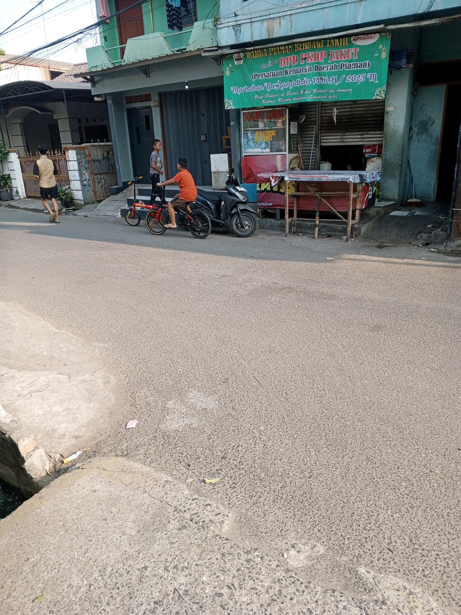 The image depicts a street scene in North Jakarta, Indonesia, on a sunny day. In the foreground, the textured asphalt road dominates, with a concrete curb and a small drain visible on the left. In the mid-ground, two young boys are present. One boy, wearing an orange t-shirt, rides a red folding bicycle on the road. The other boy, in a grey t-shirt, stands on the sidewalk near the bicycle. Behind them, a black motorcycle is parked.
Further back, a building with a light green upper story and a grey lower story is visible. A green banner with white lettering hangs across the building's facade, reading "WARGA PIAMAN BERBAGI TAKJIL GRATIS! DPD PKDP JAKUT (Persatuan Keluarga Daerah Piaman) Marhaban Ya Ramadhan 1446 H / 2025 M". Below the banner, a storefront with a closed metal gate is partially visible, with a table covered in red and white plastic sheeting in front of it. A person wearing a beige vest and black shorts walks away from the camera on the sidewalk. Electrical wires are strung across the top of the image. The overall environment suggests a residential or commercial street in a bustling Indonesian city.