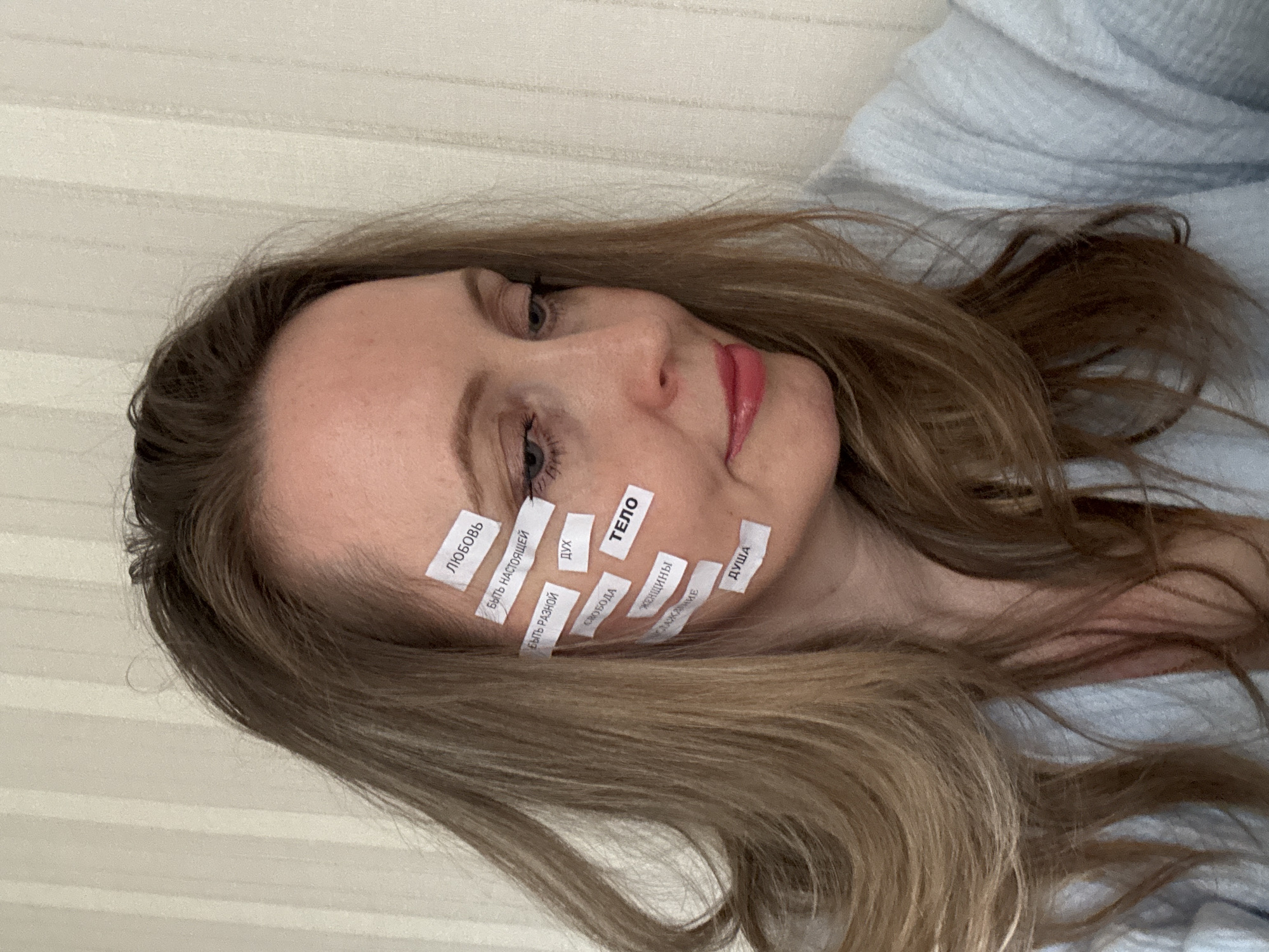 A woman with light brown hair is depicted lying down, resting her head against a light blue, textured fabric, possibly a robe or blanket. The background features a cream-colored wall with faint horizontal stripes. Her head is tilted, and her gaze is directed towards the right side of the frame (her left). She has several small, white slips of paper with Russian words printed on them, taped onto her face, cheek, and temple. Her expression appears neutral or slightly pensive.
The visible Russian words on the slips include:
* "ЛЮБОВЬ" (Love)
* "БЫТЬ НАСТОЯЩЕЙ" (To be real/authentic)
* "ДУХ" (Spirit)
* "БЫТЬ РАЗНОЙ" (To be different/varied)
* "ТЕЛО" (Body)
* "СВОБОДА" (Freedom)
* "ЖЕНЩИНЫ" (Women)
* "ИСТОРИЯ" (History/Story)
* "ДУША" (Soul)
The lighting suggests an indoor setting, likely during the day, but no specific time cues are present. The image conveys a thoughtful or introspective mood, possibly exploring themes related to self-identity and personal concepts.