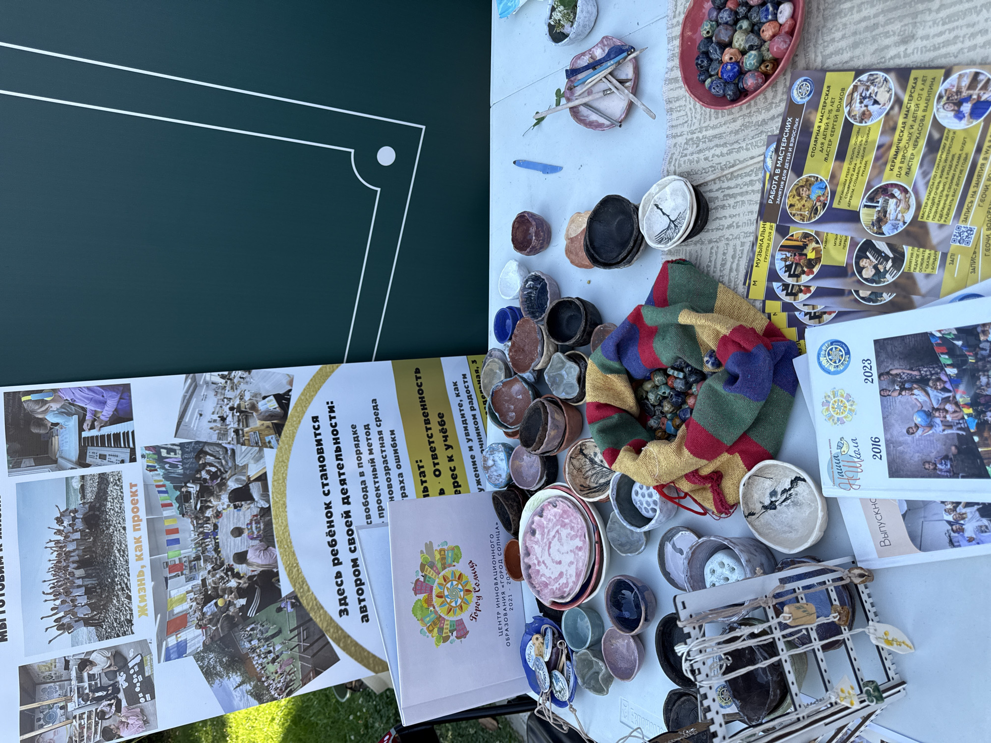 This is a medium shot, eye-level outdoor photograph taken in the city of Sochi, Russia, on a table displaying various handmade ceramic items and informational booklets. The background is dominated by a dark green surface with white geometric lines, and to the left, a large poster with images of children engaged in activities.
On the table, there are numerous small ceramic bowls and dishes of various colors and designs, including some with intricate patterns. A colorful striped fabric lies next to a collection of beads. A wooden rack holds several necklaces with ceramic pendants. Several books and brochures are also present, with one in the foreground titled "Выпускной" (Graduation) and another with the title "Школа" (School). The brochures advertise various master classes, including pottery and music.
The lighting is natural and appears to be daylight. The overall scene suggests a craft fair, exhibition, or educational event, possibly related to a school or creative center.