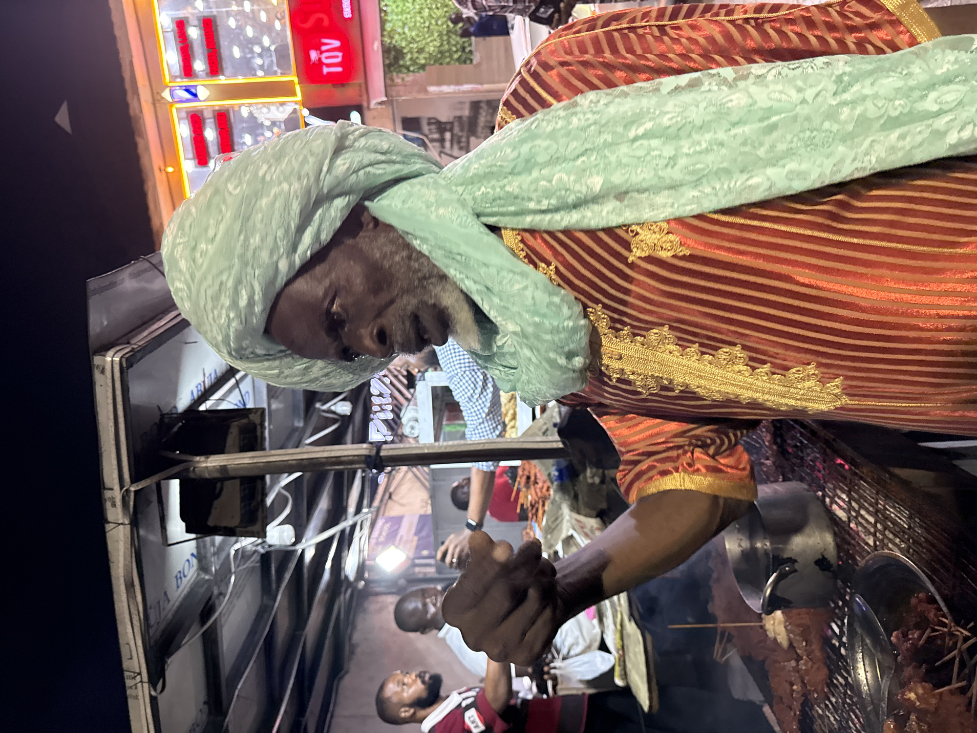 An older man wearing a traditional red and gold striped robe and a light green turban is cooking skewers of meat on a grill at a street food stall in Ado, Nigeria. He is reaching down to turn or tend to the meat. Other people are in the background, some waiting for food. The stall has a metal frame. A sign with the words "TOV STORES" is visible in the background along with other city lights.