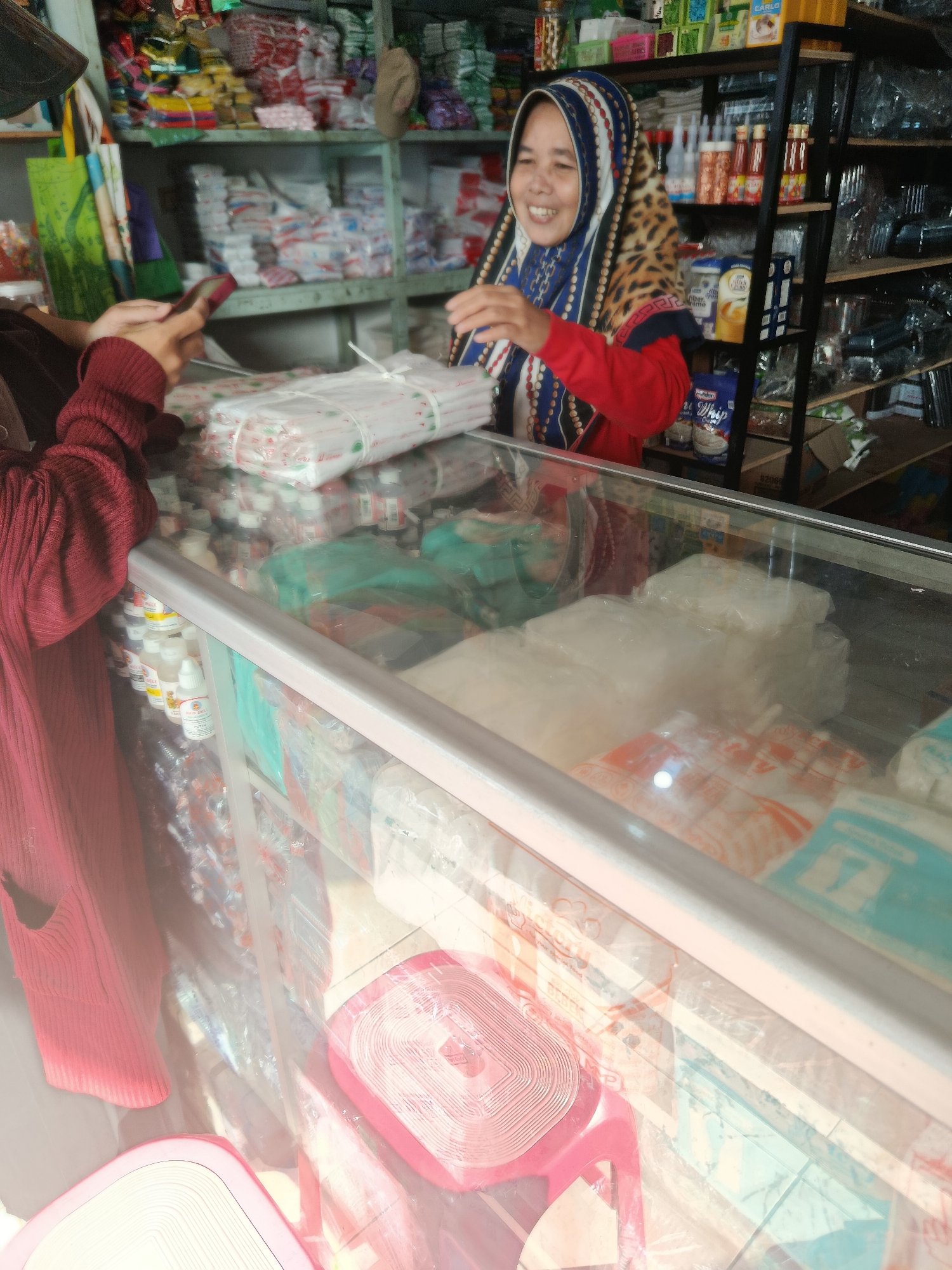 In Kalijeruk, Indonesia, a woman wearing a hijab and a red top is at a market stall, interacting with a customer whose hand is visible holding a phone. The stall is filled with various items, including packaged goods, plastic containers, and bottles on shelves behind the woman. A glass display case in front of her contains more packaged items. The overall scene depicts a commercial transaction within a bustling market environment.