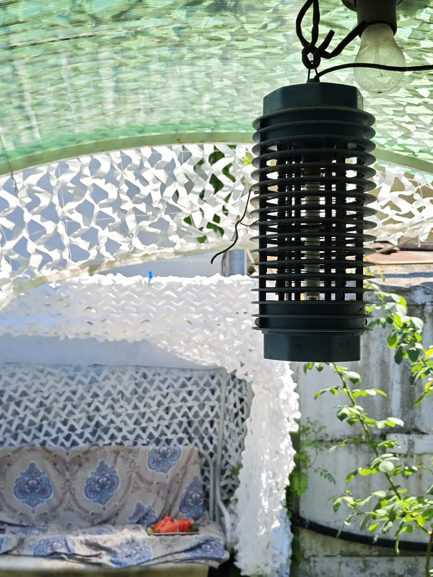 An outdoor scene in Rostov-on-Don, Russia, shows a dark cylindrical insect zapper hanging from a wire. The zapper is positioned beneath a green, mesh-like canopy and next to a vintage porch swing draped with white lace and a patterned fabric. A plate of sliced watermelon rests on the swing. Green foliage from a nearby bush is visible to the right of the zapper.