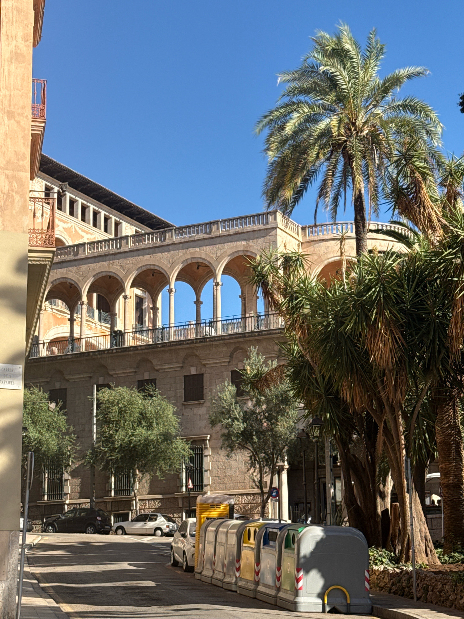 The image depicts a sun-drenched street scene in Palma, Spain, likely in a historic district, with a large, ornate building featuring a loggia with arched openings dominating the background. The building's facade is a warm, ochre hue, adorned with decorative details and balconies. Lush palm trees and other foliage frame the building and line the street, casting dappled shadows on the asphalt. Several cars are parked along the side of the road, and a row of colorful recycling bins is positioned on the sidewalk. A street sign is visible on the left side of the image, partially obscured. The sky is a clear, vibrant blue. The overall impression is one of a picturesque Mediterranean urban environment on a bright day.