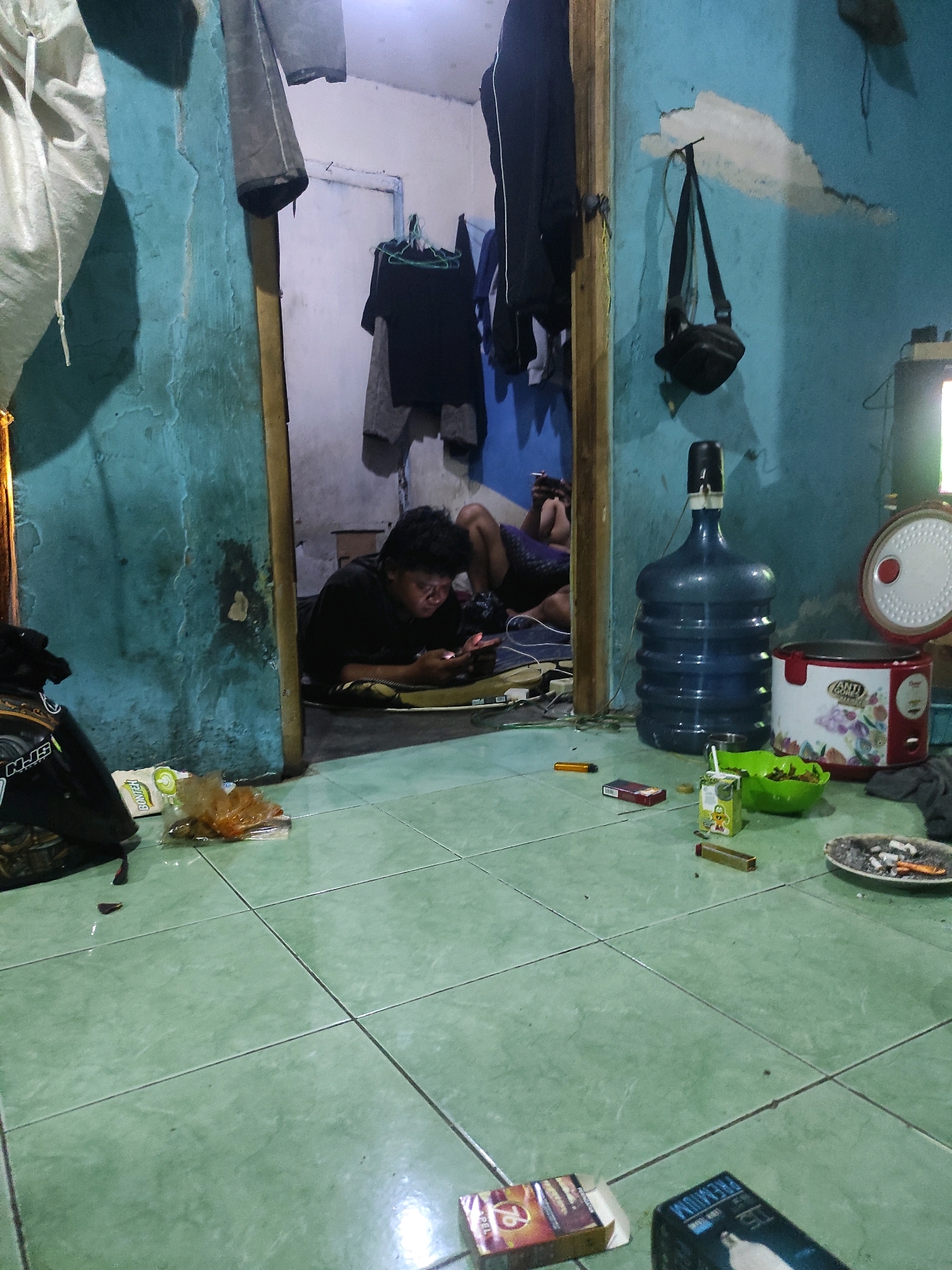 In Laksanamekar, Indonesia, a dimly lit room with distressed turquoise walls and a green tiled floor shows two young men engaged with their phones. One man, lying on his stomach on a dark mat, is deeply focused on his device. The other man, partially visible, is also interacting with a phone. The room is cluttered with various items, including a large blue water cooler bottle, a rice cooker, a green bowl with food, a cigarette pack, a lighter, and a cigarette butt in an ashtray. Clothing hangs on a rack in the background, and a black bag is slung over a hook on the wall. The overall impression is of a casual, somewhat untidy domestic interior.