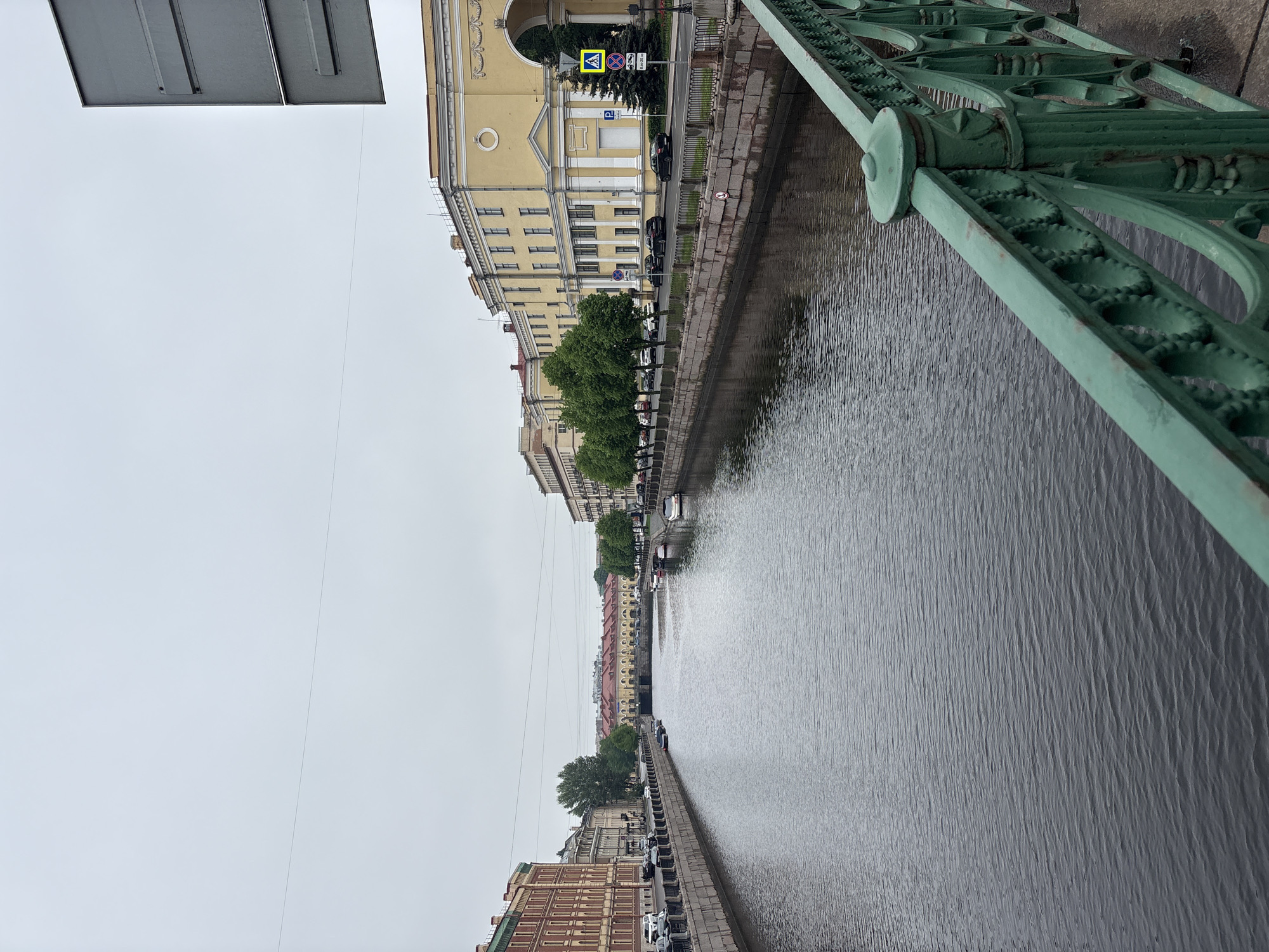 This is an eye-level, full outdoor shot of a canal in Saint Petersburg, Russia, viewed from a bridge. The canal reflects the overcast sky and the buildings lining both sides. The bridge has a green, ornate railing visible on the right. Buildings of varying heights and architectural styles, primarily yellow and brick, line the canal banks. Some trees are present along the embankment. Cars are parked along the edge of the canal, and road signs are visible.