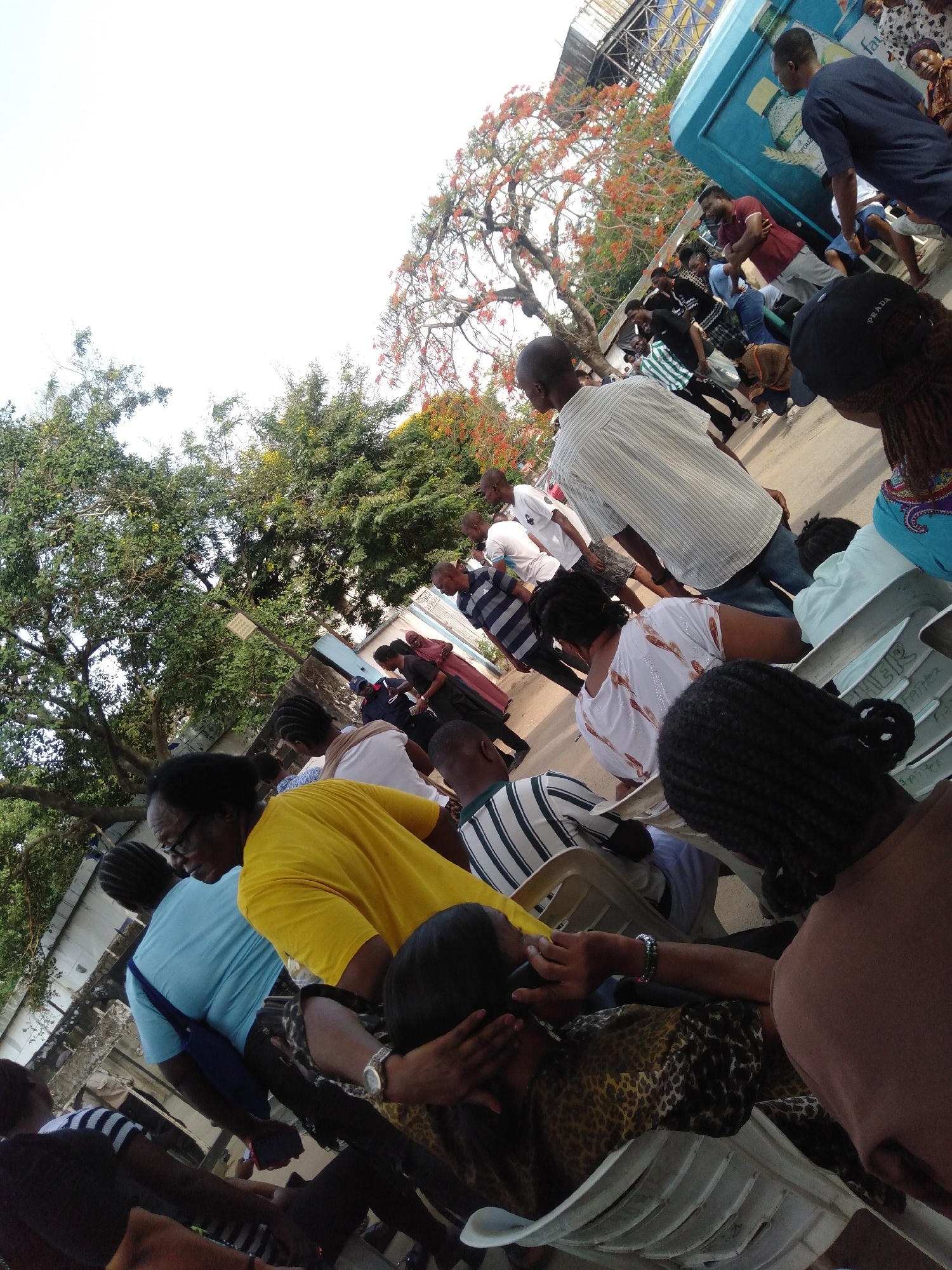 The image captures a scene in Lagos, Nigeria, showing a gathering of people outdoors. Many are seated on white plastic chairs, suggesting an organized event or waiting area. A group of people are standing, with some appearing to be walking or moving around. There's a mix of clothing styles and colors, including yellow, blue, and patterned garments. Trees with green and reddish-orange foliage are present. A cooler or service vehicle is visible in the background, along with some infrastructure. The overall impression is that of a public event or a waiting area with people milling about.