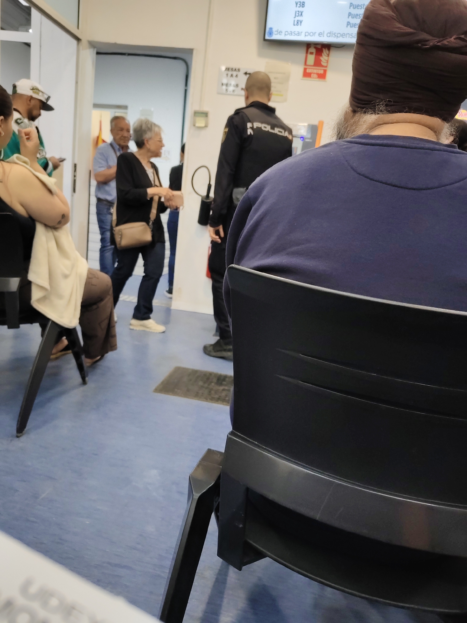 The image depicts an indoor scene within a public administrative office or waiting area in Montcada, Spain. The environment features a blue linoleum floor and white walls, with an open doorway in the mid-ground.
In the immediate foreground, a person is seated with their back to the camera, wearing a dark brown turban and a dark blue long-sleeved top, showing a visible grey beard. They occupy a black plastic chair. To their left, another individual is partially visible, seated in a black chair, wearing a light-colored top, brown pants, and sandals, with a beige cloth draped over their arm.
In the mid-ground, a police officer, identifiable by "POLICIA" on their dark uniform, stands facing away from the camera, positioned near a wall-mounted digital screen. To the officer's left, an older woman with short grey hair and glasses is seen in profile, wearing a black cardigan, dark pants, and light shoes, holding a light brown shoulder bag. She appears to be interacting with a man in a light blue shirt and jeans who is partially visible behind her. Further left, a man in a white baseball cap and a green and white jacket can be seen holding a phone.
The wall-mounted screen displays various alphanumeric codes like "Y3B," "J3X," "L8Y," and the partial word "Puest" (likely "Puesto," meaning station or counter). Below these, part of the Spanish phrase "de pasar por el dispensa" is visible, suggesting instructions for passing through a dispensary or service point. A red sign with "EXTINTOR" (fire extinguisher) is also mounted on the wall. Additional text from OCR includes "JESAS," "144," "VEAS," "5-6," and the partially obscured "UDE" in the bottom left. The scene conveys a typical daytime atmosphere in a public service facility where people are waiting or moving between areas.