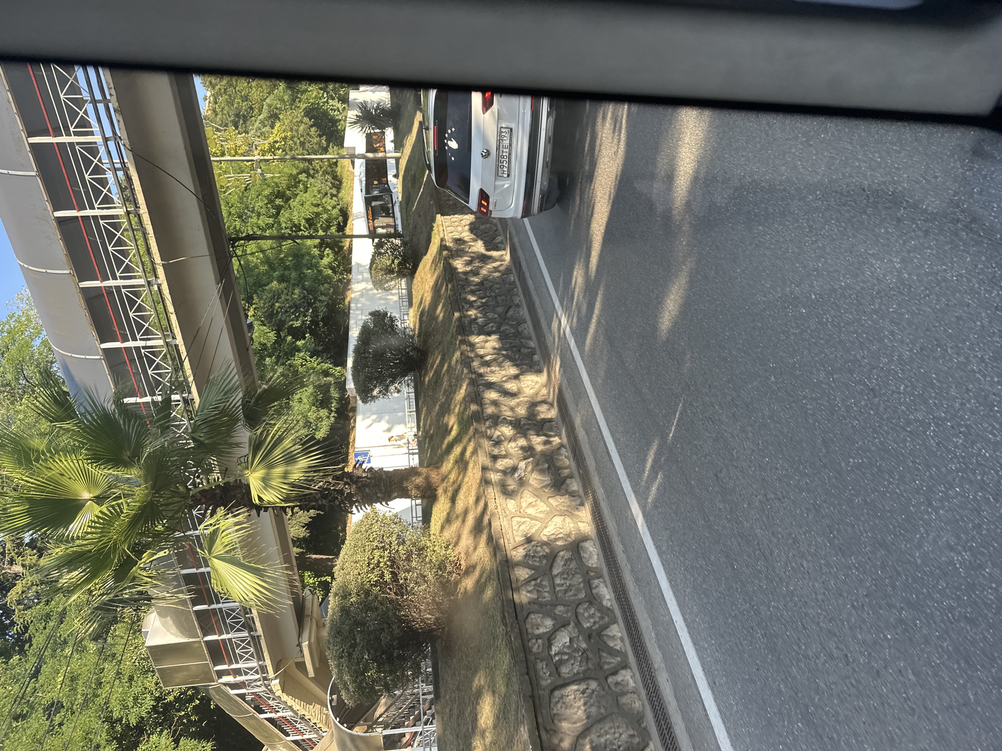 The image, taken from inside a vehicle, depicts a street scene in Sochi, Russia, characterized by a pedestrian overpass and lush greenery. A white Volkswagen SUV, with license plate H958TE 193, is the prominent vehicle in the foreground, driving on a paved road. The SUV has a decorative decal on its rear window, a circle with several star-like shapes. Behind the SUV, a bus is partially visible, suggesting public transportation is present. The overpass, a significant architectural element, spans across the road, featuring a covered walkway with white railings and red accents. The surrounding environment is rich with vegetation, including palm trees and dense green foliage, indicative of Sochi's subtropical climate. A stone retaining wall runs along the side of the road, supporting a grassy embankment dotted with bushes and trees. Sunlight casts shadows across the scene, indicating it is daytime. The image captures a typical urban landscape with elements of infrastructure and nature intertwined.