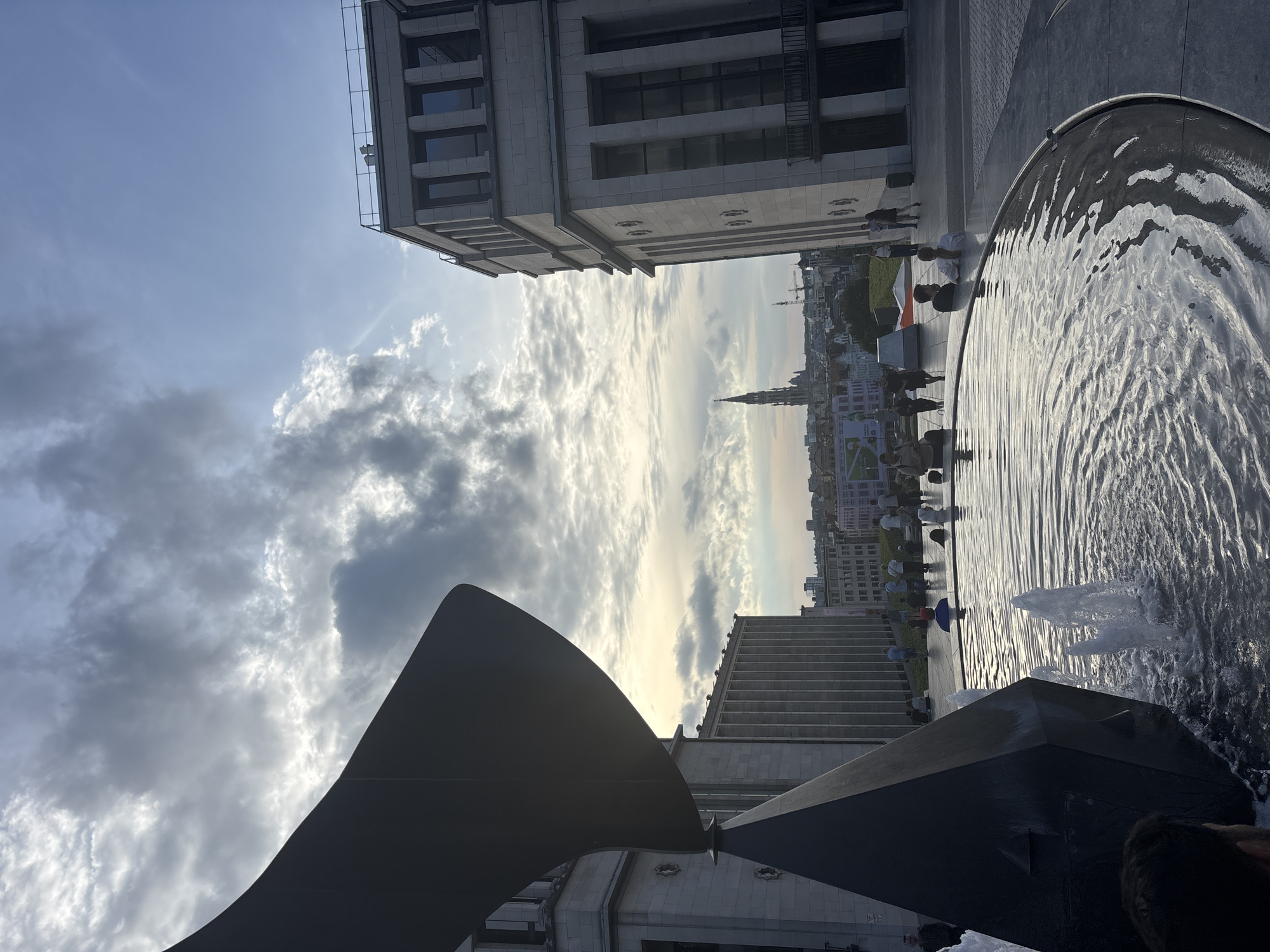 The image captures a contemporary urban scene in Brussels, Belgium, specifically Place de la Monnaie, featuring a modern fountain and surrounding architecture. In the foreground, a large, dark abstract sculpture is partially submerged in a circular fountain with water jets creating ripples and splashes. Several people are visible, some seated on the fountain's edge, others standing or walking in the background. The architectural style of the buildings is predominantly neoclassical, with the facades of two prominent structures flanking the fountain. In the distance, the cityscape of Brussels extends, with a prominent church spire rising above the rooftops, suggesting a historical contrast to the modern elements in the foreground. The sky is partly cloudy, with soft, diffused light filtering through, creating a slightly overcast atmosphere. The overall impression is one of a public space where modern art and architecture coexist with the historical urban fabric.