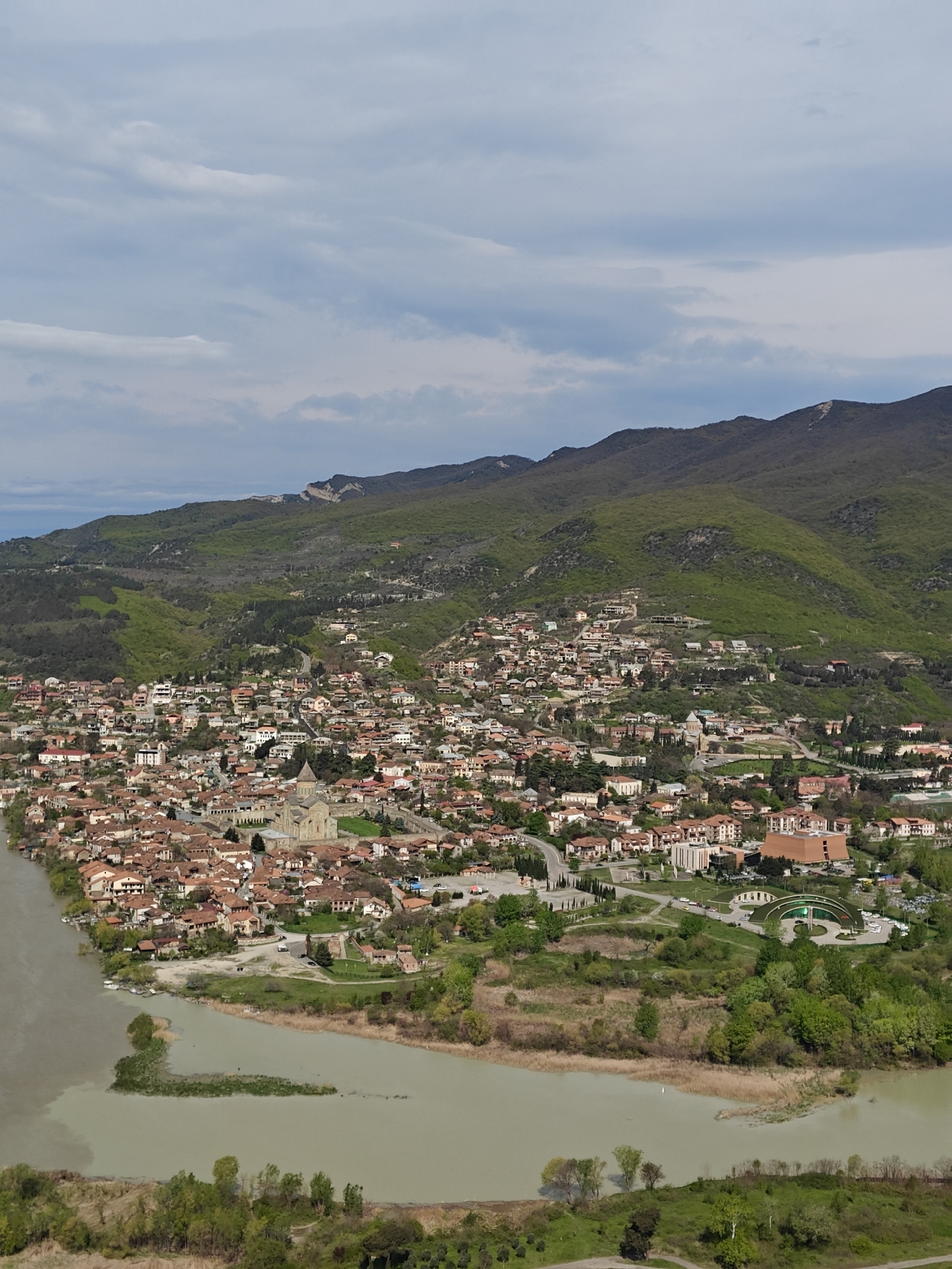 The image shows an aerial view of a city nestled near the confluence of two rivers, set against a backdrop of green, hilly terrain and a partly cloudy sky. The city features numerous buildings with reddish-brown roofs, clustered together with a prominent cathedral-like structure at the center. A network of roads and pathways meanders through the urban landscape. The rivers are wide and muddy in color, with some vegetation and small islands visible near the riverbanks. The hills in the background are covered with trees and vegetation, with some exposed rock faces visible. The sky is a mix of blue and white, with scattered clouds. Overall, the scene depicts a peaceful and picturesque urban environment surrounded by natural elements.