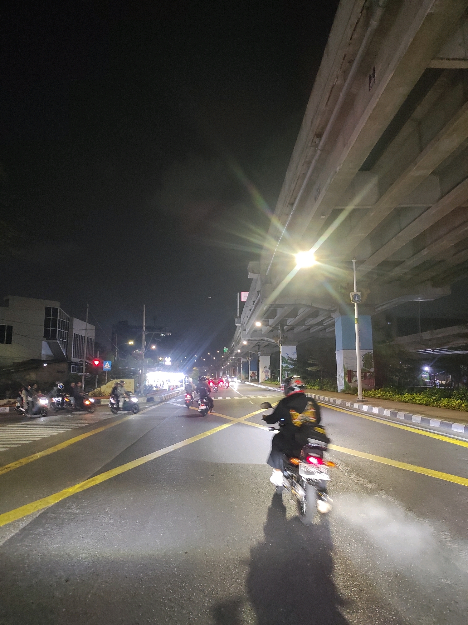 The image depicts a street scene at night in East Jakarta, Indonesia, likely near an overpass or elevated structure. The foreground is dominated by a paved road with distinct yellow lane markings. A person is riding a motorcycle away from the camera, emitting a plume of exhaust. The motorcycle's taillight is visible, and a shadow of the rider and bike is cast on the road. In the midground, several other motorcycles are visible, some with riders, traveling in the same direction. On the left side of the road, there are buildings and a pedestrian crossing with a traffic light showing red. The right side of the road features a sidewalk with a row of plants and streetlights illuminating the scene. The background is dark, with the silhouette of an elevated concrete structure overhead. The overall atmosphere is that of a busy urban street at night, with traffic and illuminated commercial areas.
