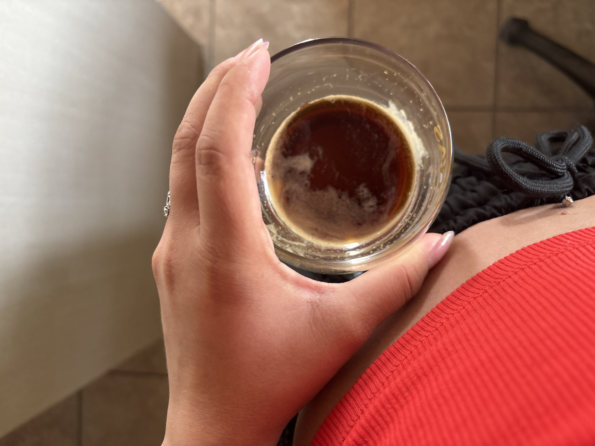 A close-up, high-angle shot captures a person holding a clear glass containing a dark brown beverage, likely coffee, with a layer of light foam or crema on top. The subject's hand, with a light skin tone and manicured nails featuring a light polish, is prominent in the center-left, gently gripping the glass. A silver-colored ring with a small, clear gemstone is visible on the ring finger.
The person is wearing a vibrant red, ribbed fabric garment, partially visible on the right side of the frame, and a black garment with a drawstring, which appears to be shorts or sweatpants, just below the glass.
The background, partially visible on the left and upper right, consists of neutral-toned tiled surfaces, possibly a floor and a lower wall section, indicating an indoor setting. A blurry dark object is present in the upper right background. The image focuses tightly on the hand and drink, with the overall scene being well-lit. The activity depicted is simply holding a drink. No specific time of day or emotions are discernible, and no text is visible in the image. The scene is located in Orizari, Bulgaria.