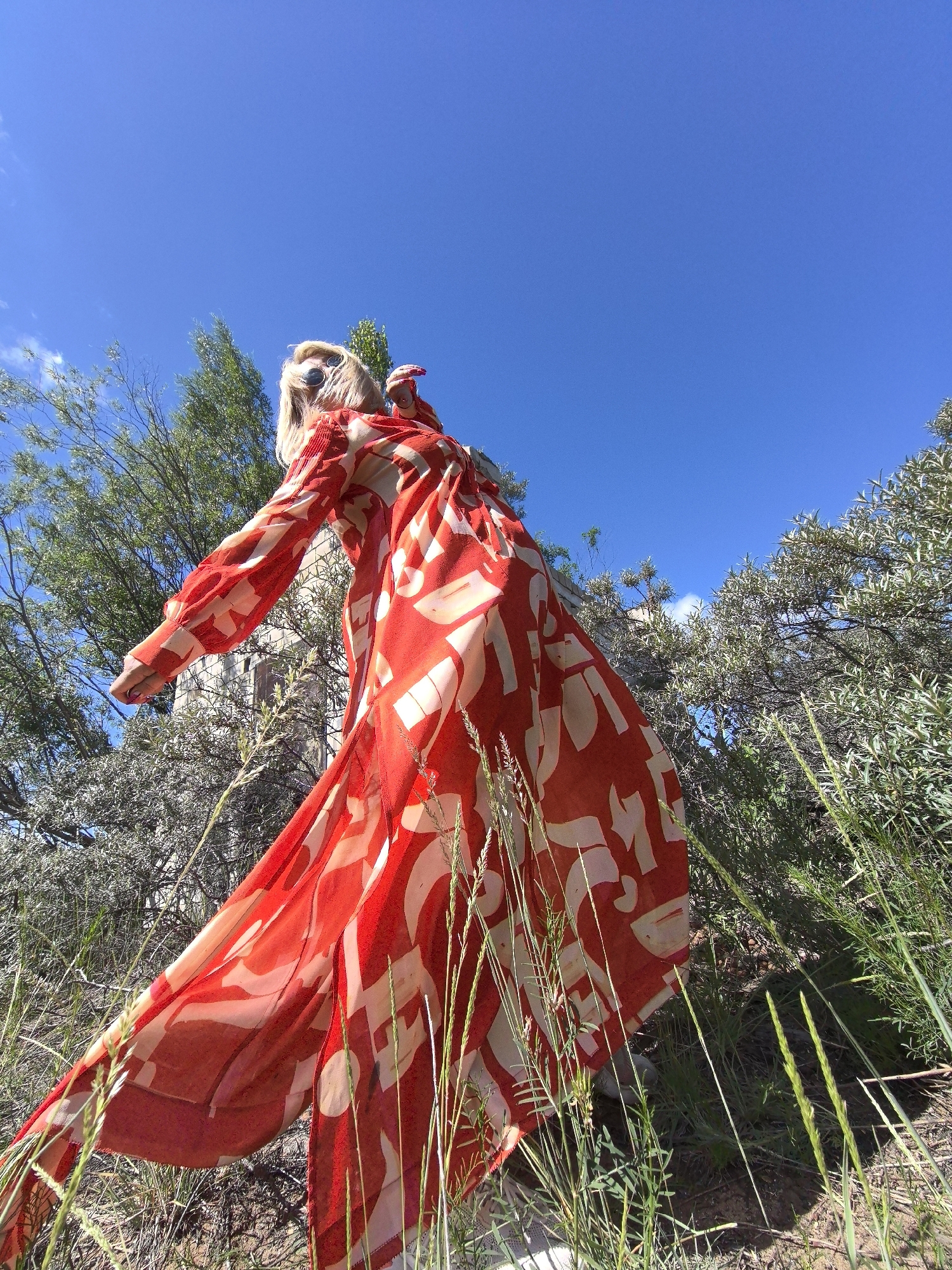 A woman with blonde hair and sunglasses is standing outdoors, looking up at the sky. She is wearing a long, flowing red and white patterned dress. The woman is positioned in the foreground, with her back to the camera and her body angled to the left. Behind her, there is foliage and a stone structure, possibly a building or wall, which is out of focus. The background is a clear blue sky. The scene is bathed in bright sunlight, creating strong shadows. The image appears to be taken from a low angle, looking upwards. The location is not specified as Noviy, Russia; there is no visual information to support this claim.