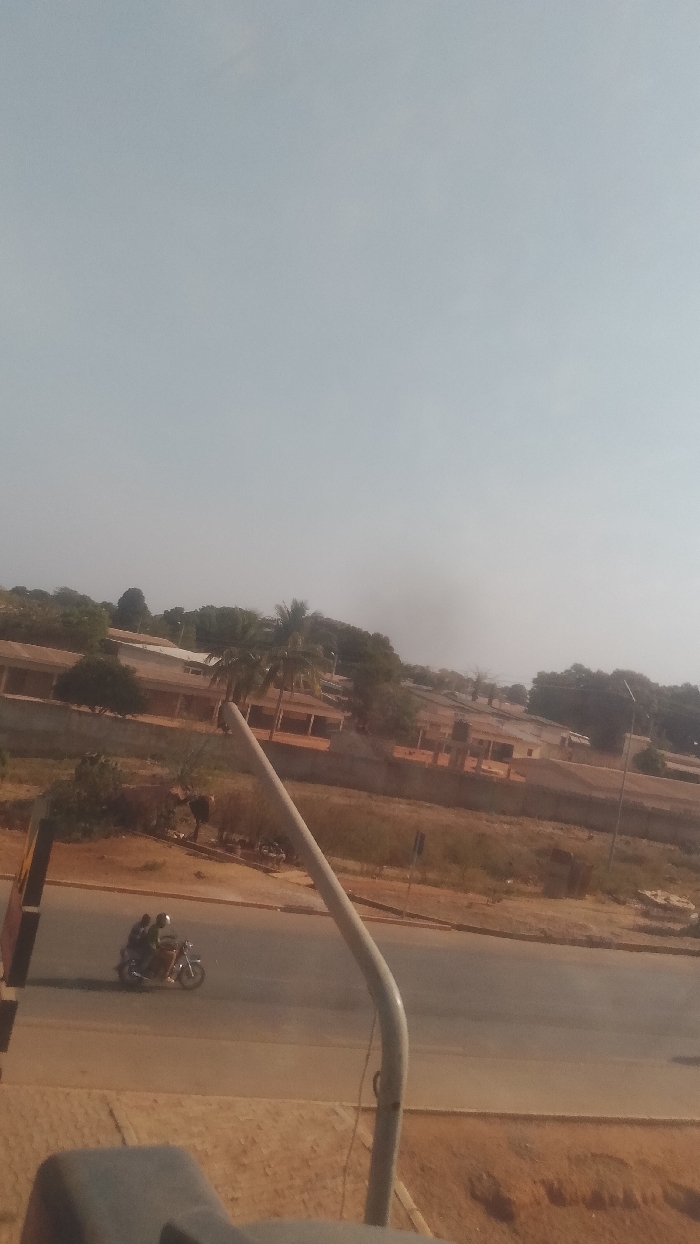 The image presents an elevated view of an urban landscape in Korhogo, Côte d'Ivoire, under a light, hazy blue-grey sky. A faint, dark plume of smoke or haze is visible near the horizon. In the immediate foreground, a dark, indistinct object partially occupies the lower-left, next to a curved, light grey metal pole extending diagonally from the lower-right towards the upper-left. Beneath the pole, a reddish-brown dirt verge transitions to a two-lane paved road. On this road, a white motorcycle travels from left to right. The motorcycle carries two individuals: the driver, wearing a light-colored top and a dark helmet, and a passenger seated behind, wearing a dark top and a dark helmet. Their shadows are cast on the pavement. Across the road, a wide expanse of reddish-brown earth, featuring sparse dry vegetation, precedes a series of single-story buildings. These structures possess light-colored walls, darker roofs, and open portico-style fronts. Numerous trees, including several tall palm trees and other dense foliage, are interspersed among the buildings and extend towards the background. A utility pole with a light fixture is visible on the far right side, beyond the road.