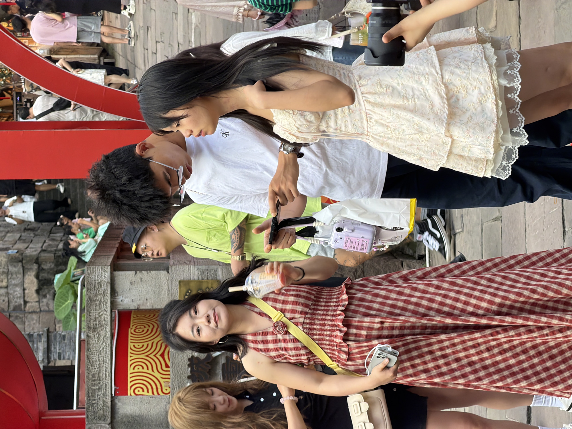 In the city of Yuzhong District, China, a group of young adults are gathered on a street. In the foreground, a woman in a red and white checkered dress holds a cup and a smartphone. To her right, a man in a white t-shirt looks down at a phone, and next to him, a woman in a white lace dress stands with her hand on her neck. In the background, other people are walking by, some carrying bags and cameras. The scene appears to be a lively, urban environment with traditional Chinese architectural elements visible, such as the red lanterns and stone walls.