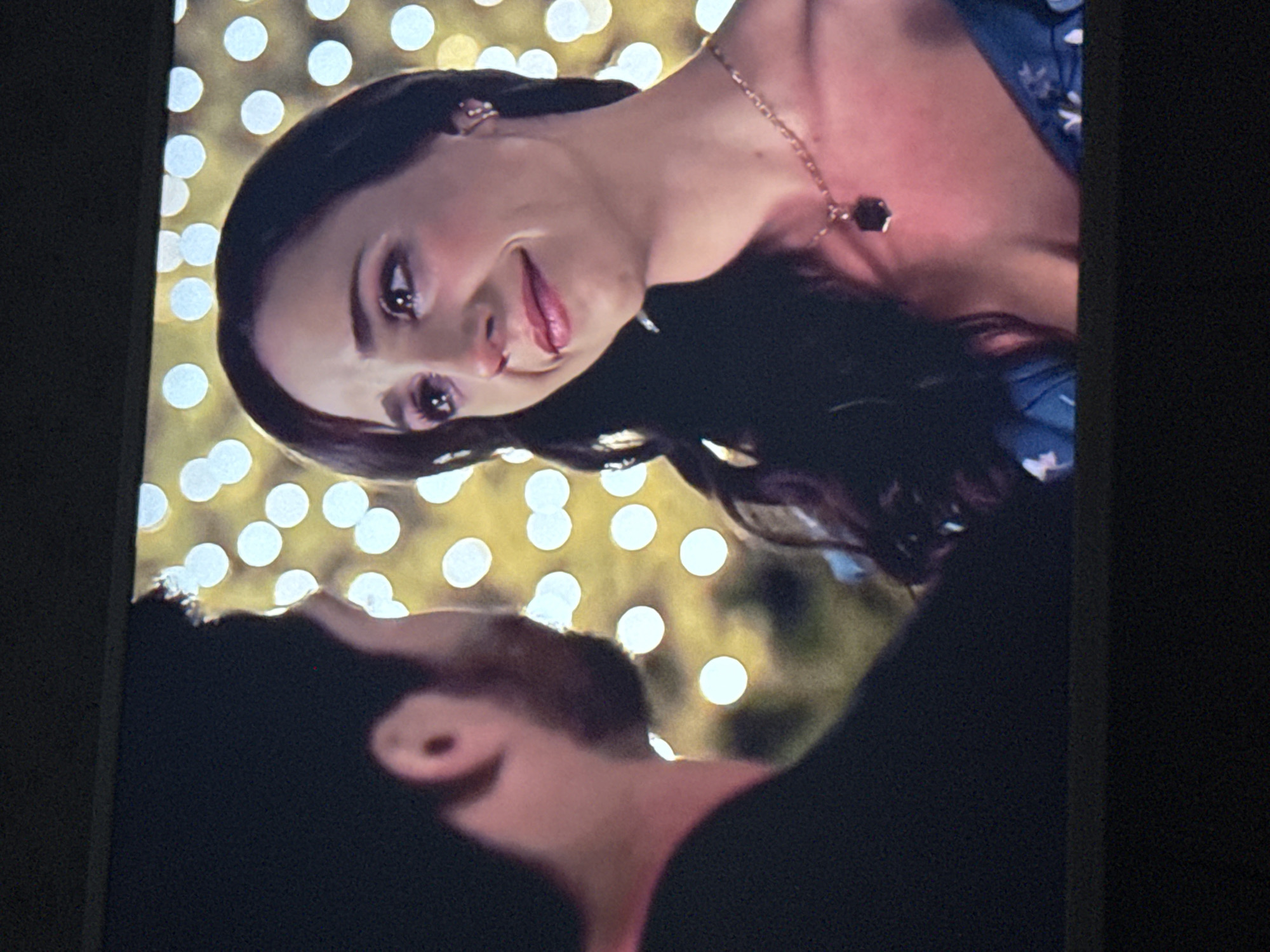 The image appears to be a photograph of a screen displaying a scene of a romantic moment. A woman with dark hair, wearing a necklace, is smiling and looking downwards at a man whose head is slightly out of focus. The background consists of numerous out-of-focus lights. There is not enough information to determine the location as Mytishchi, Russia.