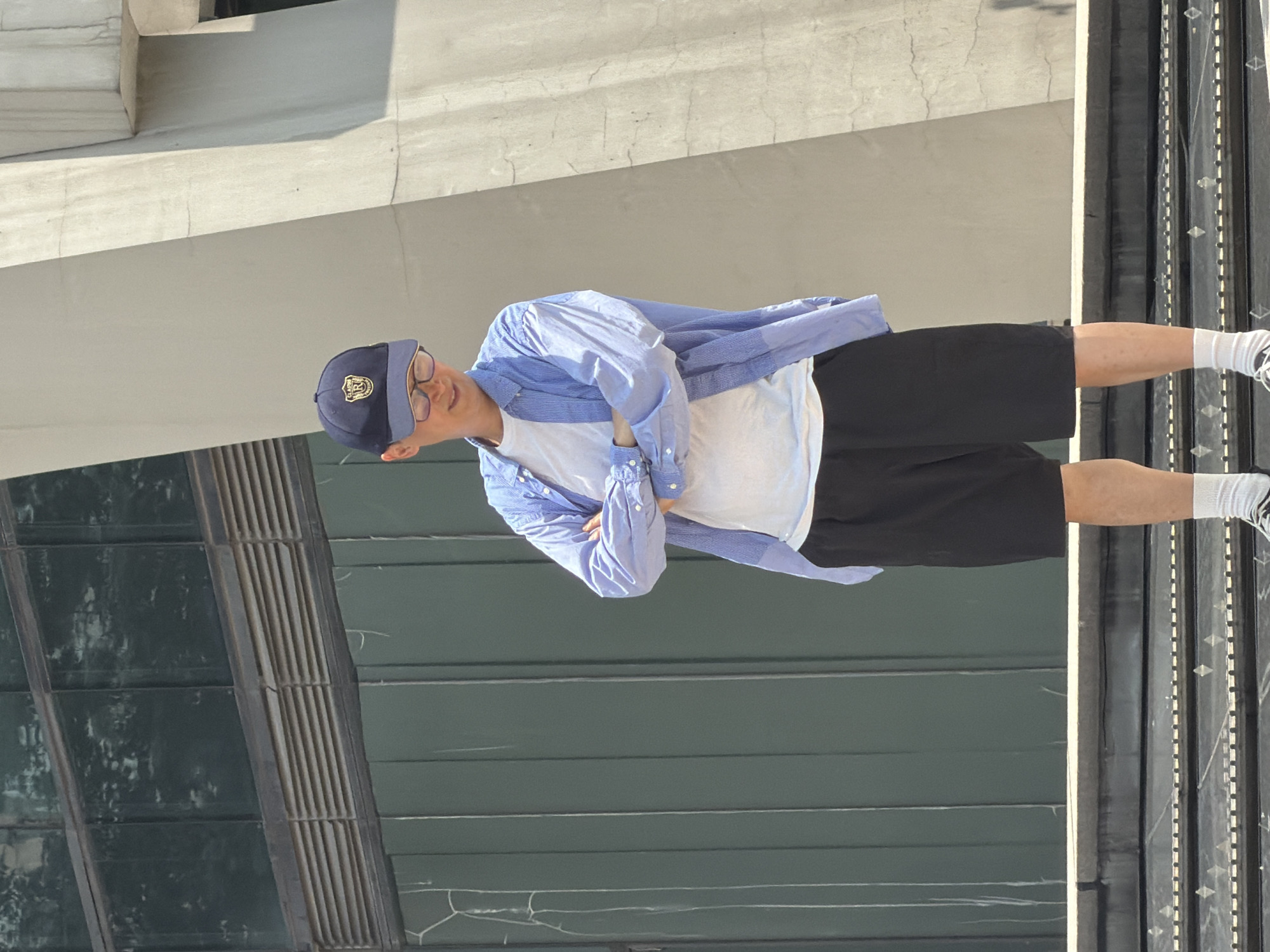 A young Asian male stands with his arms crossed on a concrete stairway in Yuzhong District, China. He is wearing a dark blue baseball cap with a gold emblem, glasses, a light blue button-up shirt over a white t-shirt, and black shorts. He has on white socks and dark shoes. The background features a building with dark green panels and reflective windows, as well as a large, angular concrete structure. The sun is casting shadows.