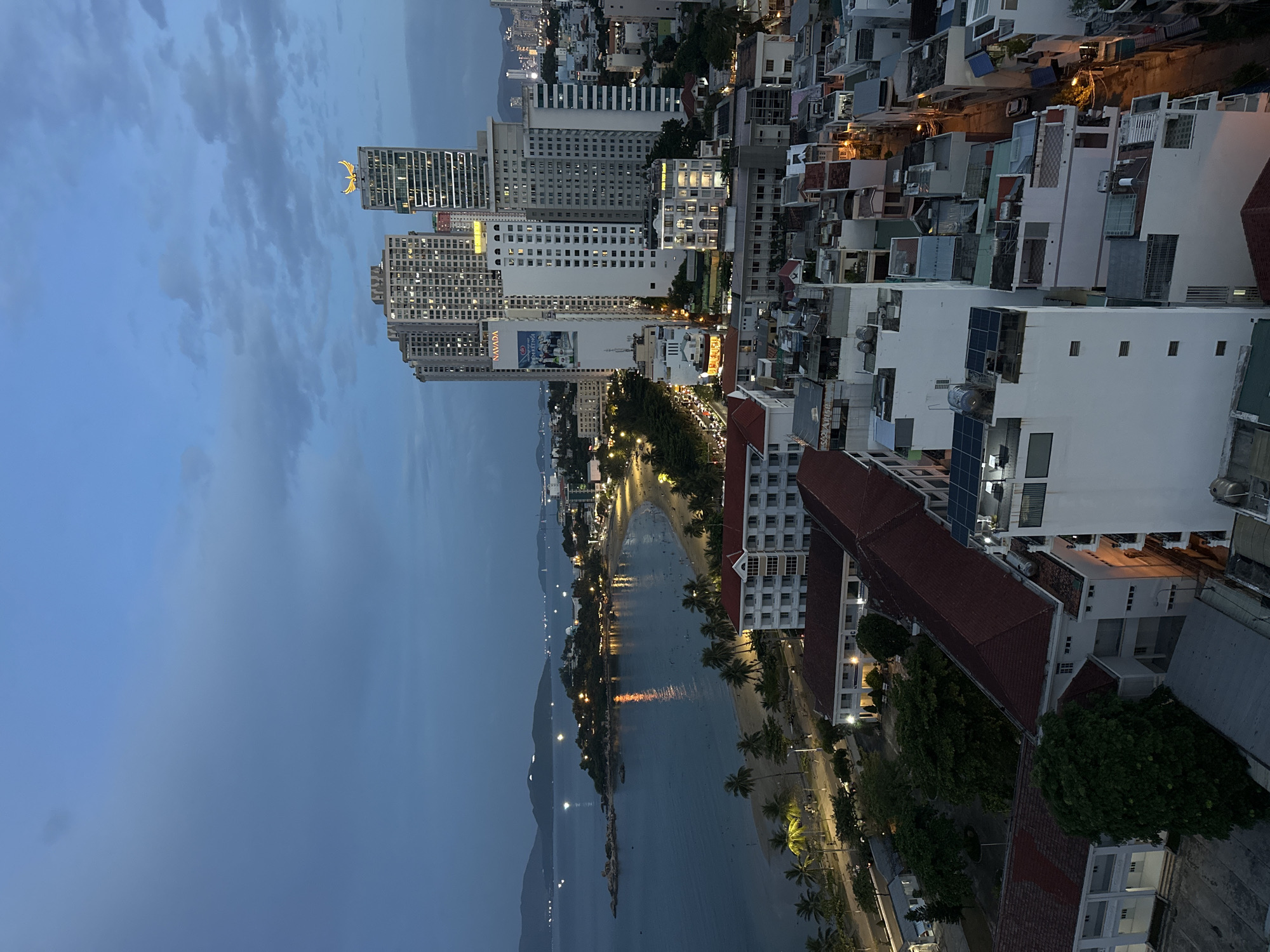 This is a twilight aerial photograph of Nha Trang, Vietnam, featuring a coastal city landscape. The image captures a wide bay with calm water, reflecting city lights. A sandy beach lined with palm trees stretches along the shoreline. Numerous buildings, ranging from multi-story apartments to smaller residential structures, dominate the urban area. Tall skyscrapers, some illuminated, are prominent in the background. The sky is a deep blue with scattered clouds. The scene depicts a bustling city transitioning from day to night, with evidence of human activity on the beach and in the illuminated streets. The overall impression is one of a coastal urban center at dusk.
