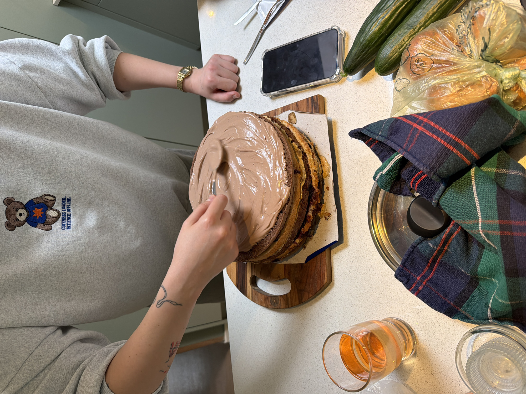 A person is in a kitchen, applying chocolate frosting to a tall, multi-layered cake. The person is wearing a grey sweatshirt with a teddy bear graphic and text that reads "CUTENESS LOADED. PATIENCE OFFLINE." They have a tattoo on their left forearm and are wearing a gold wristwatch on their right wrist. The cake is on a wooden cutting board. To the left of the cake, there is a glass of amber-colored liquid. In the foreground, there is a metal tray with a dark cloth draped over it. To the right of the cake, there is a smartphone in a clear case, two cucumbers, and a plastic bag containing round orange objects. The kitchen counter appears to be white and speckled. The background shows a glimpse of green cabinetry.