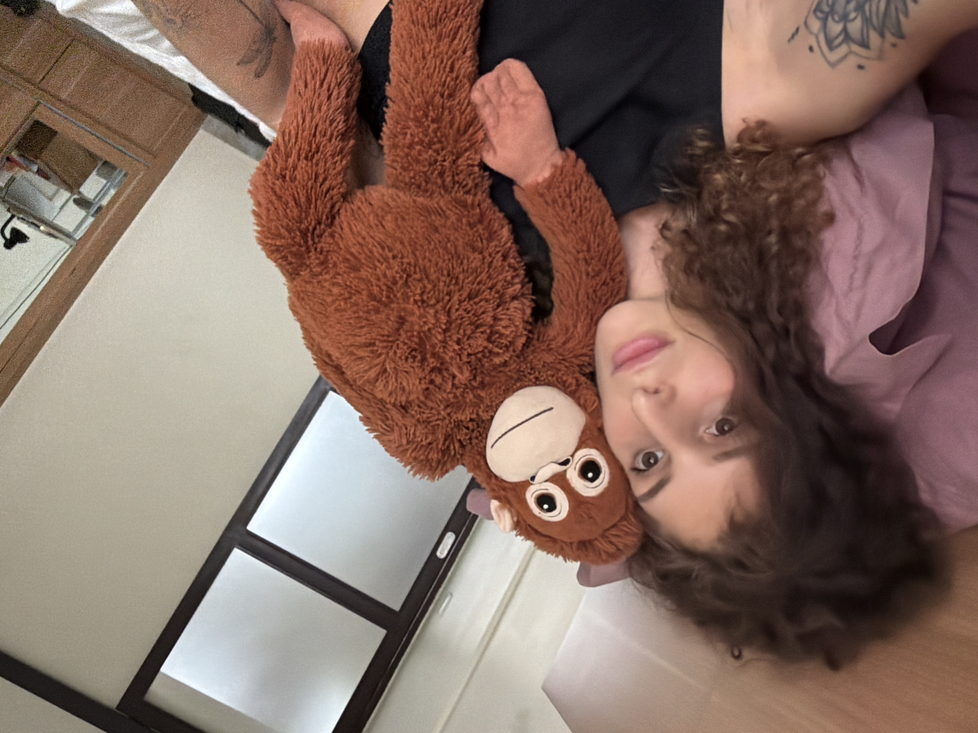 A person with curly brown hair lies on a bed in North Kuta, Indonesia, holding a stuffed orangutan. The person is wearing a black tank top and has a tattoo on their left arm. The room appears to be a bedroom, with a pink pillow visible, and a wooden dresser with a mirror in the background. A window with frosted glass and dark frames is also visible. The lighting suggests it is daytime.