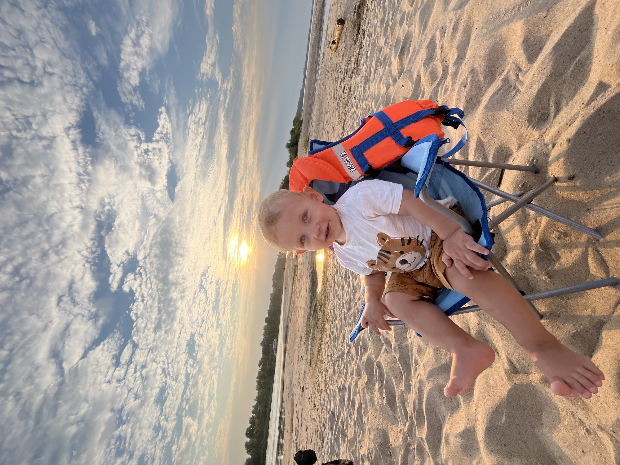 A young Caucasian child with blonde hair and blue eyes is seated in a blue and silver folding chair on a sandy beach. The child is wearing a white t-shirt with a tiger graphic and brown shorts with a tiger pattern. An orange life vest with blue straps is attached to the back of the chair. The child is facing forward, smiling slightly and looking at the camera. The background shows a wide, calm river with trees and buildings on the opposite bank. The sky is a pale blue with scattered white clouds, and the sun is setting or rising, casting a warm glow over the scene. The location is identified as the bank of the Volga River in Nizhny Novgorod, Russia.