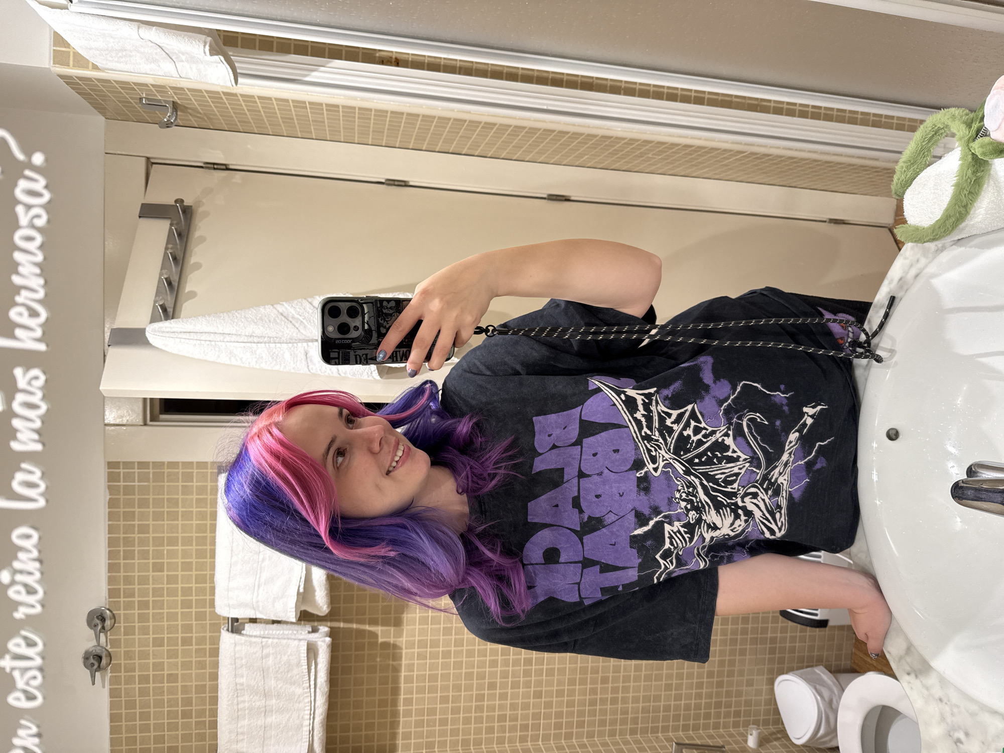 A young woman with purple and pink hair is taking a selfie in a bathroom. The bathroom features beige tiled walls, a white sink and toilet, and white towels hanging on a rack. The woman is wearing a black t-shirt with a purple "Black Sabbath" print. She is holding a black smartphone with a decorative case. The scene is likely located in a hotel or private residence bathroom, but without further information, it is impossible to confirm its location within Madrid, Spain.