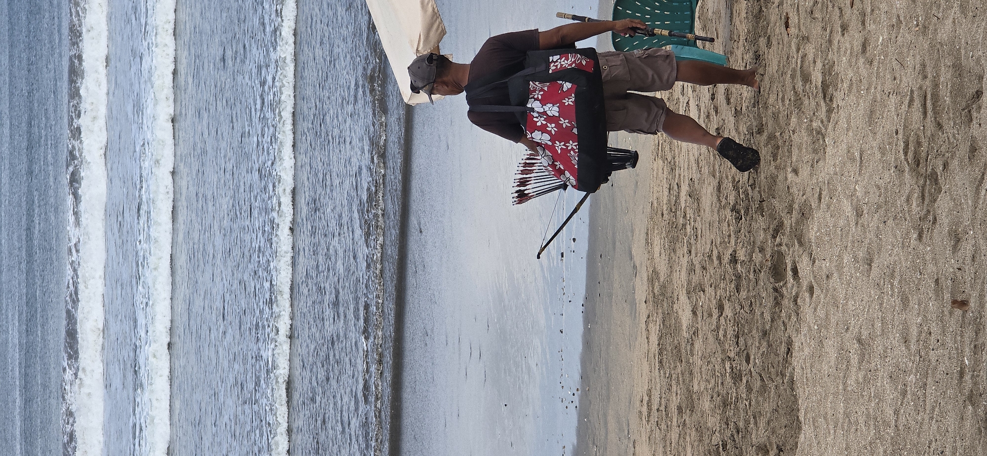 A man wearing a dark t-shirt, khaki shorts, a grey baseball cap, and black socks on his feet walks away from the camera on a sandy beach. He is carrying a large red and white floral patterned duffel bag slung over his right shoulder. The bag contains a quiver of arrows. A bow is attached to the bag, with its string taut. The man also holds a long, thin object in his right hand, possibly another bow or a staff. In the background, the ocean has several rows of waves, and the sand is littered with small debris. A part of a light-colored beach umbrella and a green plastic chair are visible to the right. The image appears to be taken in Kuta, Indonesia, known for its beaches and surf.