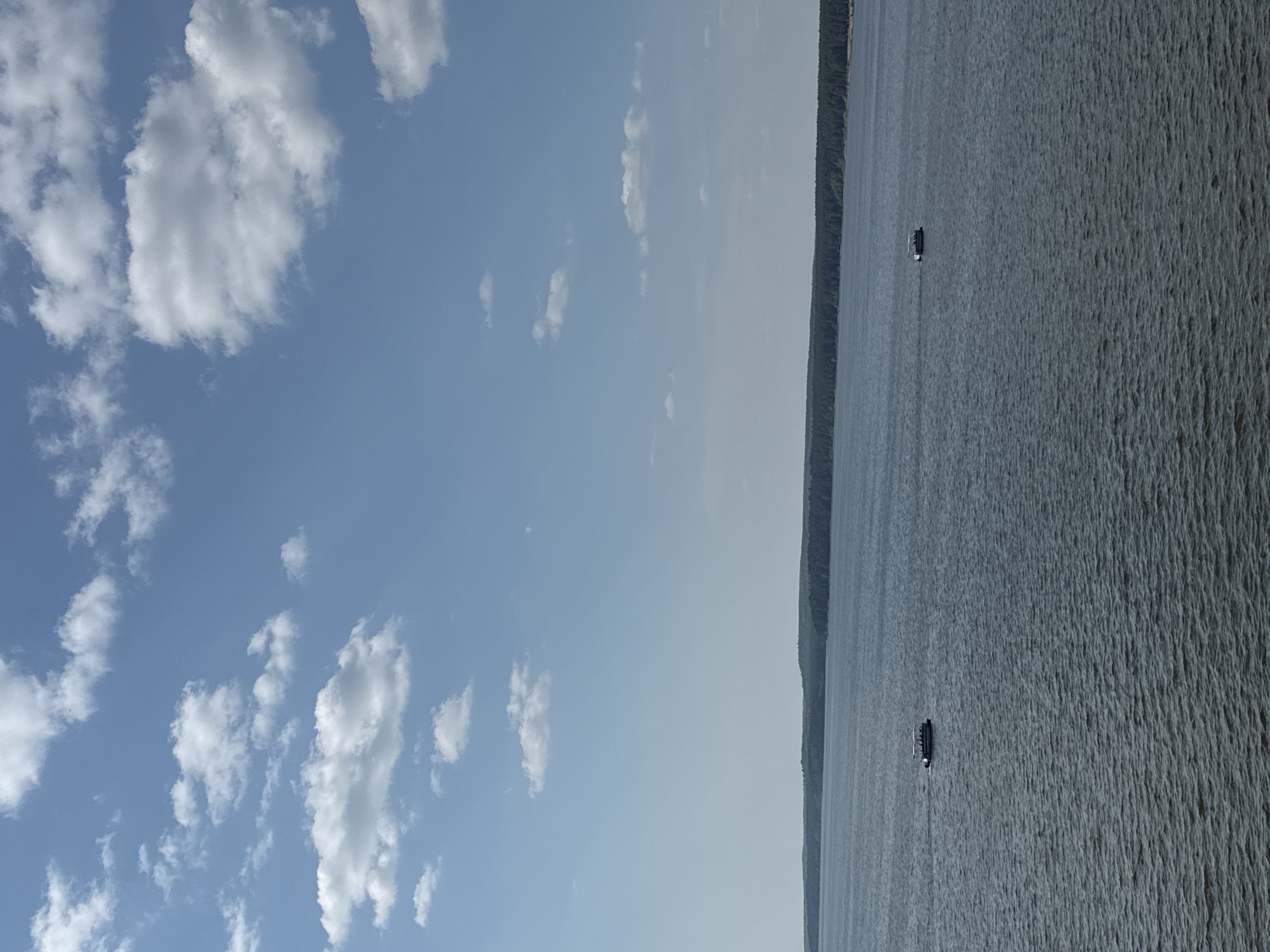 The image shows a waterscape with a clear blue sky partially covered with white clouds. The water surface has a rippled texture, suggesting a slight breeze. Two small boats are visible on the water, and a thin strip of land is seen along the horizon. Given the prompt’s reference to Votkinsk, Russia, it is plausible the scene is the Votkinsk Reservoir, located in the vicinity of the city.