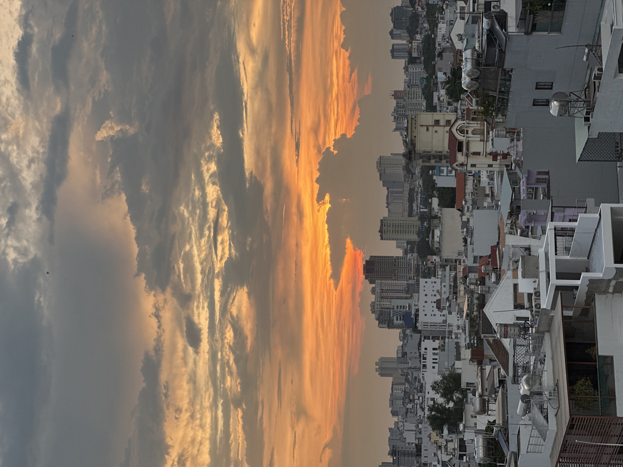 This image shows a vertical cityscape in Thủ Đức, Vietnam, at sunset. The foreground features dense clusters of buildings, primarily white and gray, with varying heights and architectural styles. The structures include residential homes, commercial buildings, and taller apartment blocks. Rooftops are cluttered with water tanks, antennas, air conditioning units, and solar panels. In the background, a vibrant sunset paints the sky with shades of orange, yellow, and gray. The clouds are striated, creating a dynamic visual effect. The lighting suggests the photo was taken during the early evening.