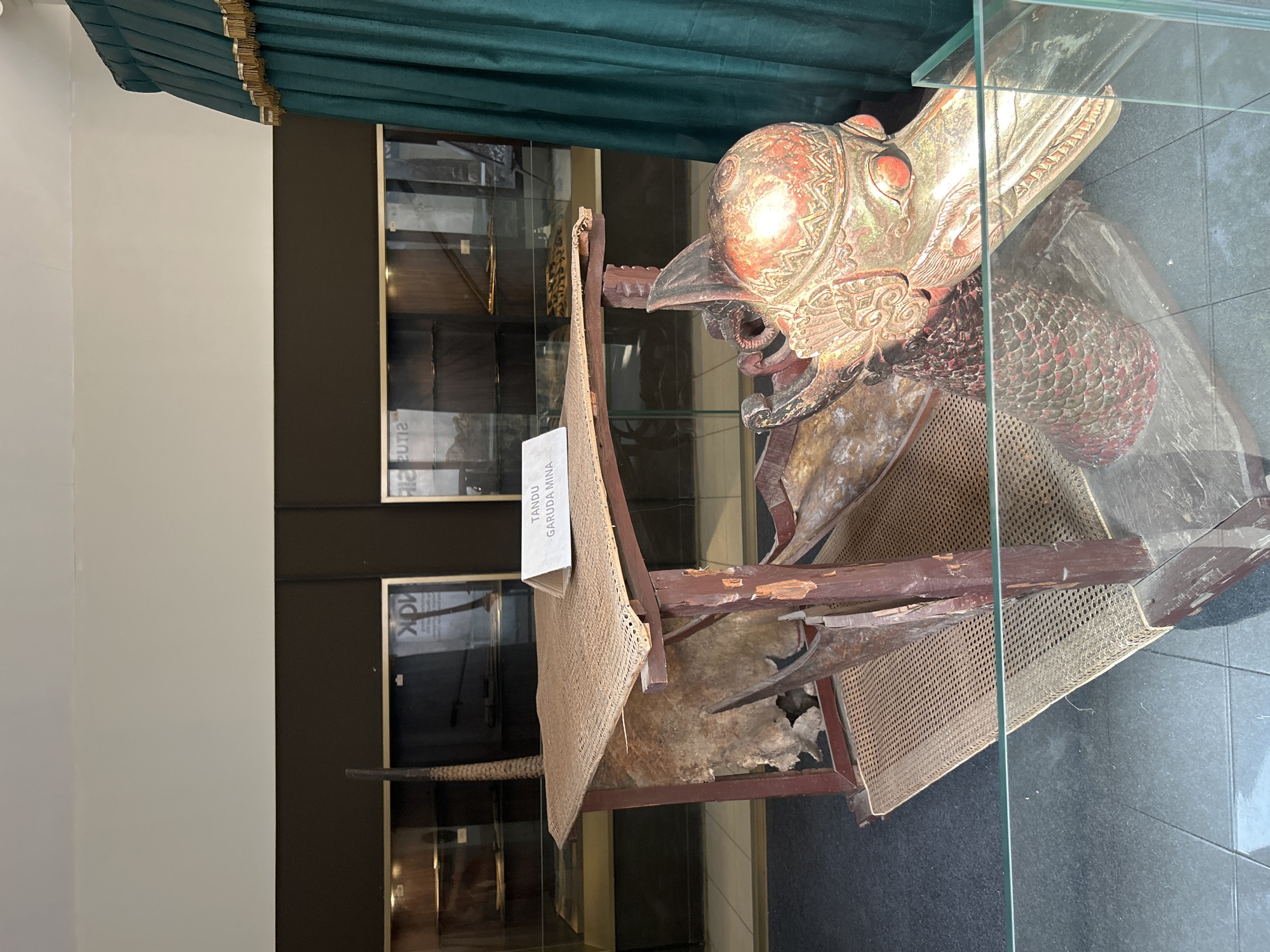 The image shows a display inside a museum. The central object is a wooden structure that appears to be a throne or a palanquin, with a woven mat for seating and a carved wooden head of a mythical creature, likely a Garuda, at the front. The creature's head is intricately decorated with red and gold paint. A sign on the seating mat reads "TANDU GARUDA MINA." In the background, display cases with various artifacts are visible, some containing what appear to be weapons or tools. A dark green curtain with gold trim hangs on the right side of the frame. The entire display is protected by a glass barrier. The context provided indicates this is located in Cirebon, Indonesia, a city known for its historical sites and cultural heritage, including its palaces and museums which often house traditional Javanese art and artifacts.