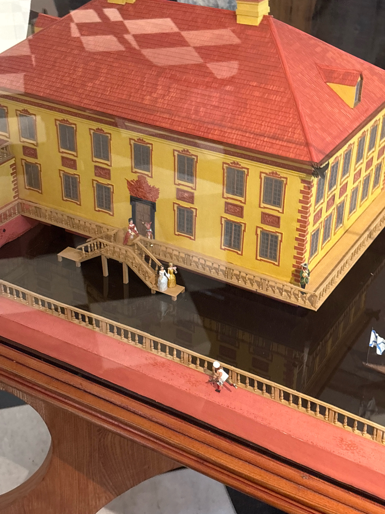 The image shows a detailed diorama of a yellow, two-story building with a red, checkered roof, resembling a historical manor or palace. The building has numerous windows with dark panes and decorative red trim. A wooden bridge with railings extends from the building over a dark body of water. Miniature figures are placed on the bridge and around the building. On the bridge, three figures in historical attire are visible, two women and one man. Near the entrance of the building, two more figures, one in a red dress and another in what appears to be a jester's costume, stand at the top of stairs. On a red walkway in the foreground, a single miniature figure in a white hat and red and white outfit is depicted. A small flag with blue and white stripes is visible in the upper right corner of the diorama. The diorama is displayed on a wooden base. The overall scene evokes a historical setting. Based on the architectural style and the presence of a flag resembling the naval ensign of the Russian Empire, this diorama could potentially represent a location related to Russian history, possibly in Saint Petersburg, Russia. However, the specific location cannot be identified from the image alone.