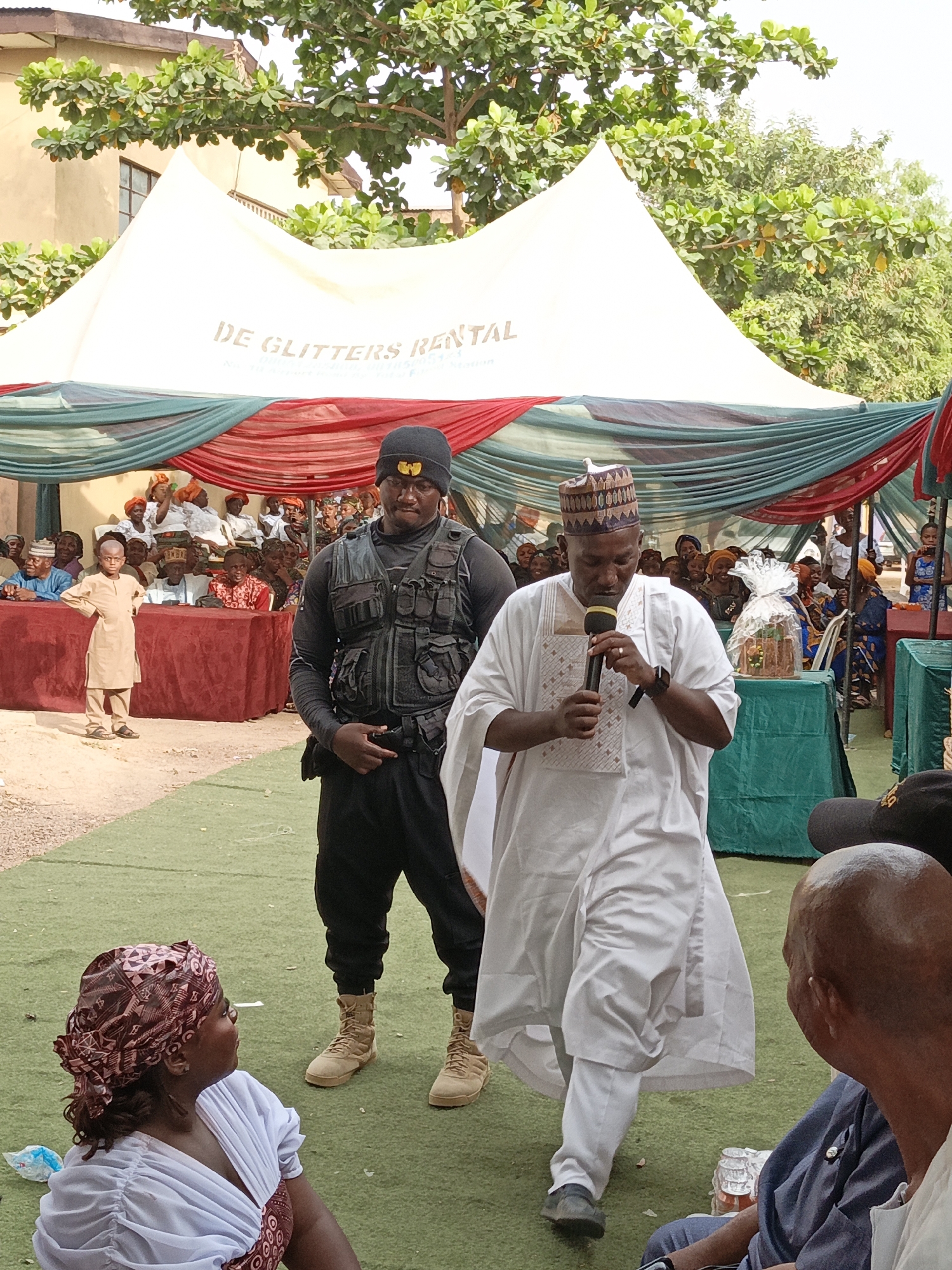 This image captures an outdoor community event taking place in Gawuna, Nigeria, under a large white tent. The tent is decorated with draped red and green fabric and clearly displays the text "DE GLITTERS RENTAL" on its side, with some unreadable smaller text below it.
In the center, a man dressed in a white traditional Nigerian tunic (agbada-like) and a patterned cap with a bird ornament is addressing an audience, speaking into a black microphone. He appears to be a key speaker or figure at the event. Beside him, slightly to his left, stands a stern-faced man, likely security personnel. He is wearing a black long-sleeved shirt, a black tactical vest with numerous pouches, black pants, and tan combat boots. A black beanie with a yellow Wu-Tang Clan logo completes his attire.
The audience consists of many people gathered for the event. In the background left, attendees are seated at tables covered with red fabric, while on the right, tables are draped in green. A young boy in a light brown traditional outfit stands near the red tables, observing the scene. Many women in the audience wear colorful headwraps. In the immediate foreground, a woman with a patterned red headwrap looks towards the speaker, and a bald man in a blue shirt is also visible. On one of the green-draped tables, a gift or cake wrapped in clear cellophane and ribbon is present.
The ground is covered with green artificial turf in the foreground, transitioning to bare earth or sand further back. Bright daylight illuminates the scene, suggesting it is daytime. Trees and buildings are visible beyond the tent structure, providing a backdrop to the gathering. The overall atmosphere points to a formal community event or celebration.