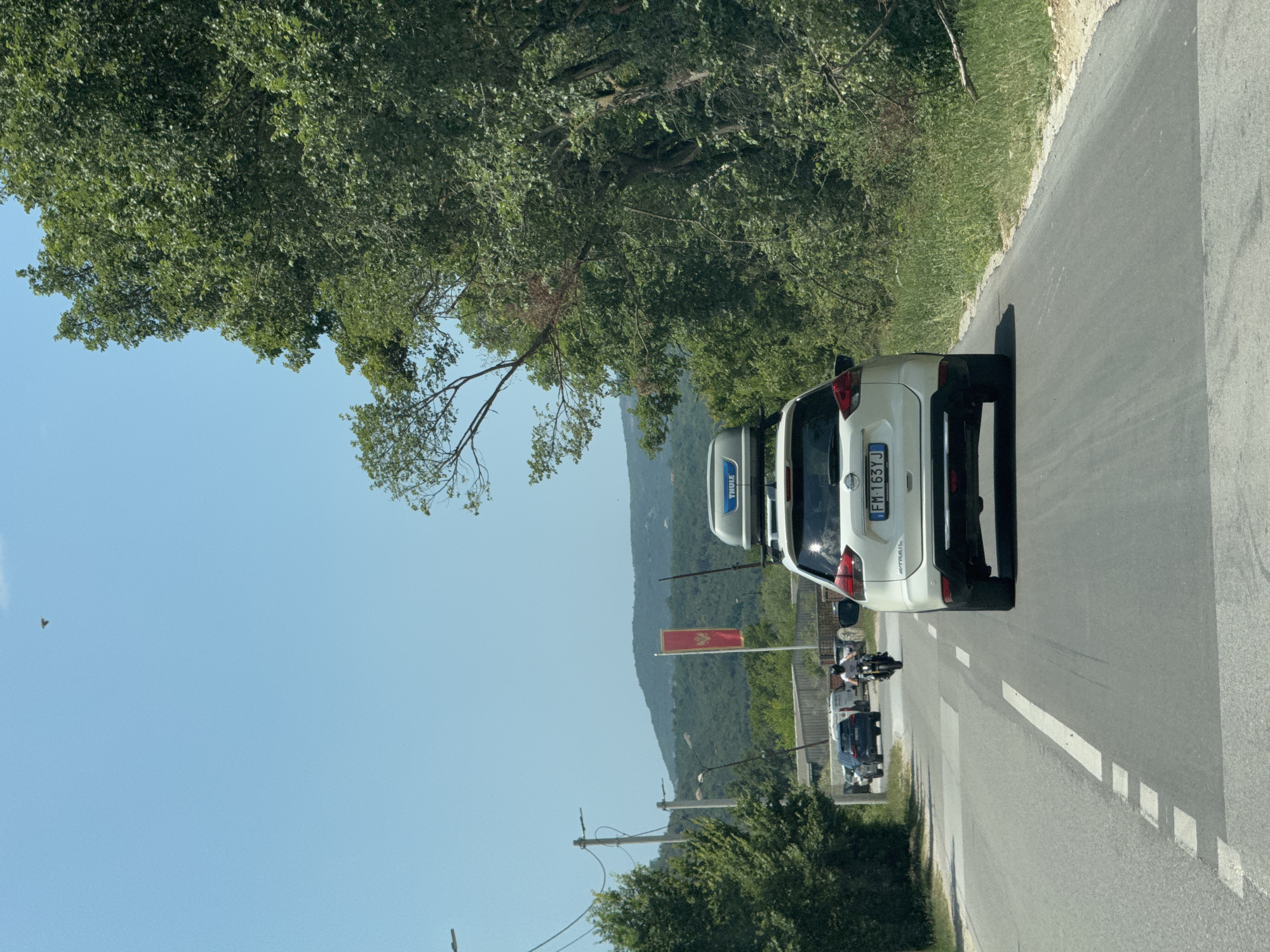 The image captures a sunny day in Đuraševići, Montenegro, with a clear blue sky overhead. The scene is a paved road bordered by lush green trees and vegetation, with rolling hills in the background.
In the foreground, a white car is parked on the right side of the road, facing away from the camera. Its license plate reads "FM-163VJ". Next to it, another vehicle, possibly a van or camper, is visible. Further down the road, several other vehicles and a person on a motorcycle are present, suggesting light traffic or a gathering. A red flag with a crest is prominently displayed on a pole to the left of the road, adding a touch of local color. The overall impression is one of a scenic, rural setting bathed in bright daylight.