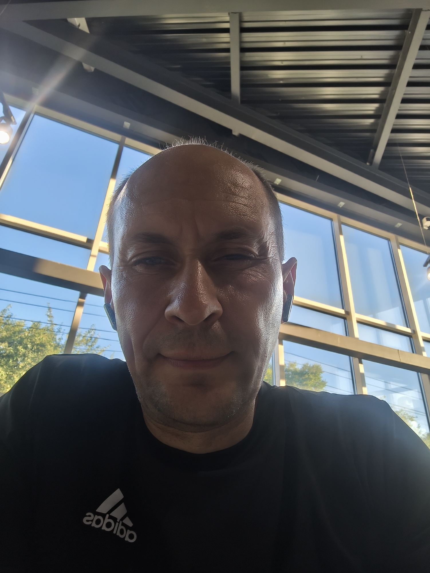 An individual is captured in a close-up, over-the-shoulder selfie, positioned indoors with a large window in the background. The window reveals a clear blue sky and the foliage of trees, suggesting an outdoor view. The interior space appears to have a modern architectural design, with exposed metal beams and panels forming the ceiling. The person, facing the camera, has short hair, is wearing earbuds, and is dressed in a black t-shirt with a white Adidas logo. The lighting suggests it is daytime, with sunlight casting reflections on the window and the person's face. There are no other individuals or objects clearly visible within the immediate frame that would allow for specific identification of a location within Komsomolsk-on-Amur, Russia.