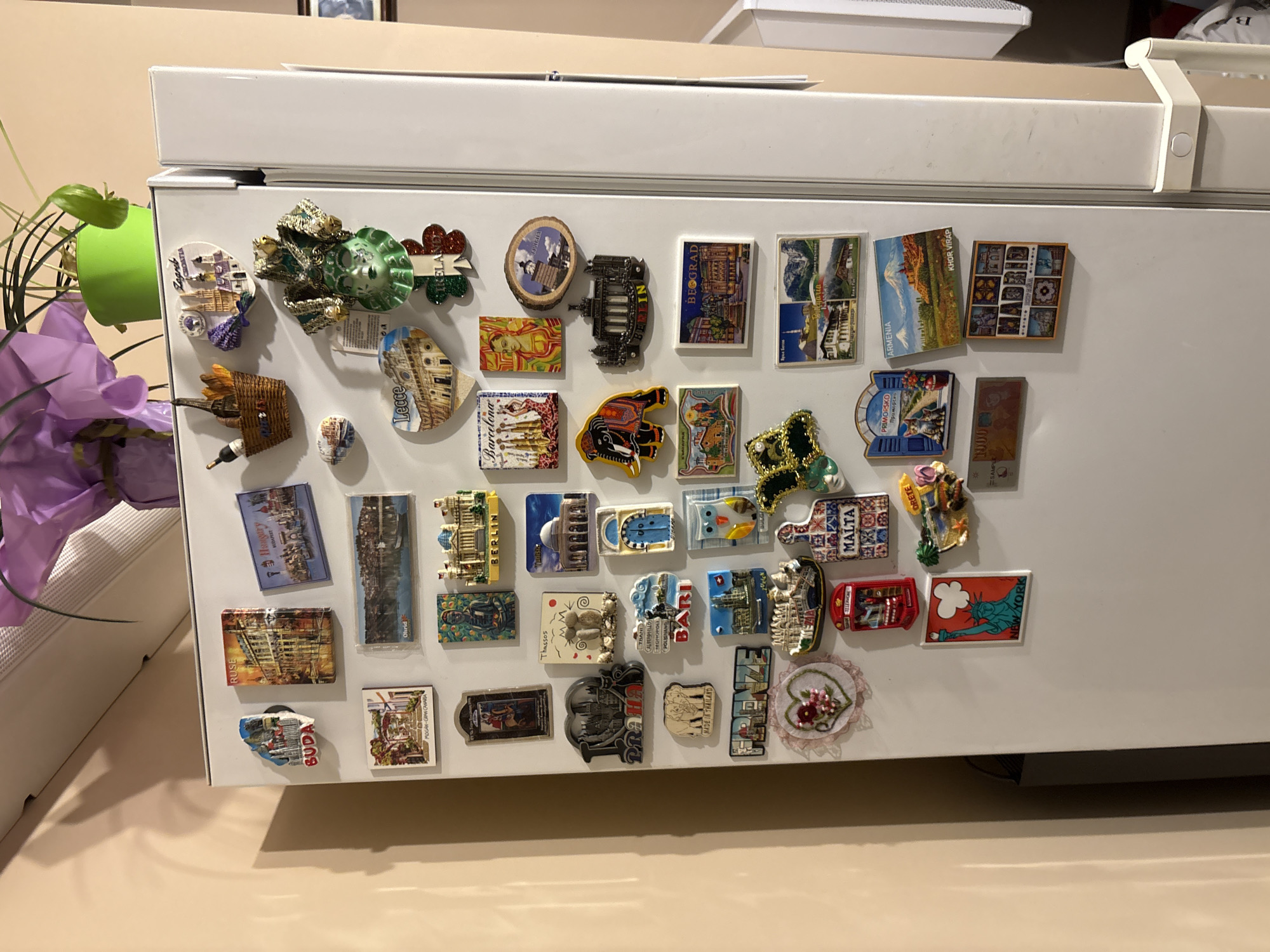 The image shows a white refrigerator door covered with various souvenir magnets. The magnets depict different cities, countries, and landmarks from around the world, including: Zagreb, Lecce, Berlin, Barcelona, Tunisia, Bari, Thailand, Malta, Crete, Armenia, Serbia, and KHOR VIRAP. The city of Sofia, Bulgaria is not depicted in the image. A purple flower is visible in the left part of the image. The refrigerator is located inside a room.