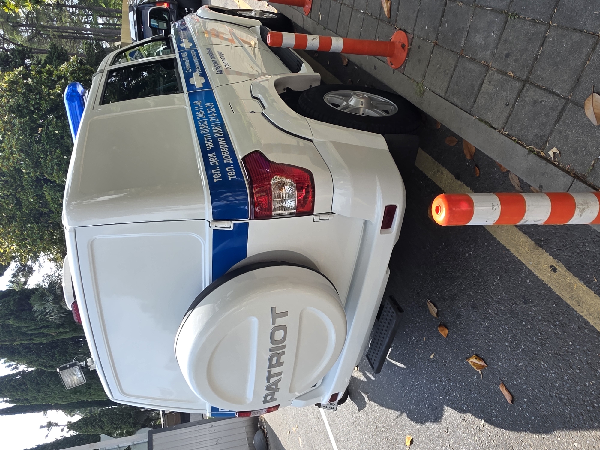 This is a portrait-oriented, full shot of the rear and side of a white official vehicle, possibly a police or emergency service van, parked outdoors. The vehicle has a blue stripe along its side and a blue light bar on its roof. The spare tire cover on the rear door is white with the word "PATRIOT" embossed in silver. Visible text on the side of the vehicle, in Russian, indicates it's related to the "Sochi PU MED" and "Adler linear police department," with contact phone numbers for the duty part and a trust line. The vehicle is parked on asphalt next to a sidewalk with a yellow line marking. Orange and white traffic cones are placed along the curb, one of which has a red illuminated top, suggesting it might be evening or late afternoon. Fallen leaves are scattered on the ground, indicating an autumn setting. The background shows lush green trees and foliage, with a hint of a darker vehicle behind the white van. The scene appears to be a daytime shot, likely in sunny weather, given the shadows. The overall impression is of a functional, official vehicle present at a specific location, possibly for operational purposes.