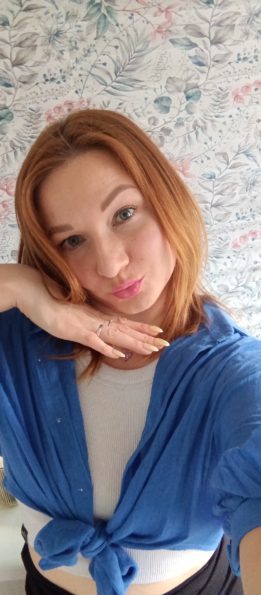A medium indoor shot features a woman with red hair, light skin, and green eyes, positioned against a wallpapered background of floral and botanical prints in muted tones. She is wearing a white ribbed tank top and a loose, draped blue shirt tied in a knot at the bottom. Her left hand is raised to her chin, showcasing two silver rings and long, gold-colored nails. A heart-shaped pendant is visible on a necklace around her neck. Her lips are pursed in a kiss. The location appears to be inside a private residence. The city is unknown, as it cannot be determined from the visual information provided.