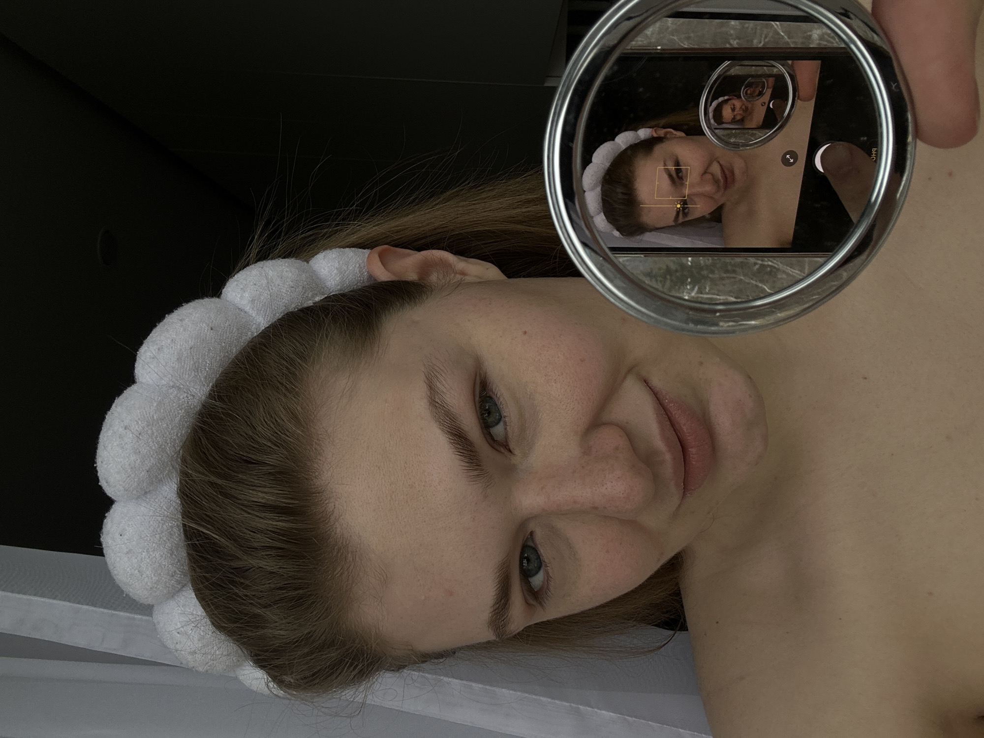A woman with fair skin, light brown hair, and blue eyes is photographed at close range, wearing a white terrycloth headband. She is holding a cell phone in her hand, using it to create a mirror effect with a round, silver-colored compact mirror. The cell phone screen displays an image of the woman herself, creating a recursive image effect. The environment appears to be an indoor setting with dark wall in the background. The location is specified as the city of Kazan, Russia.