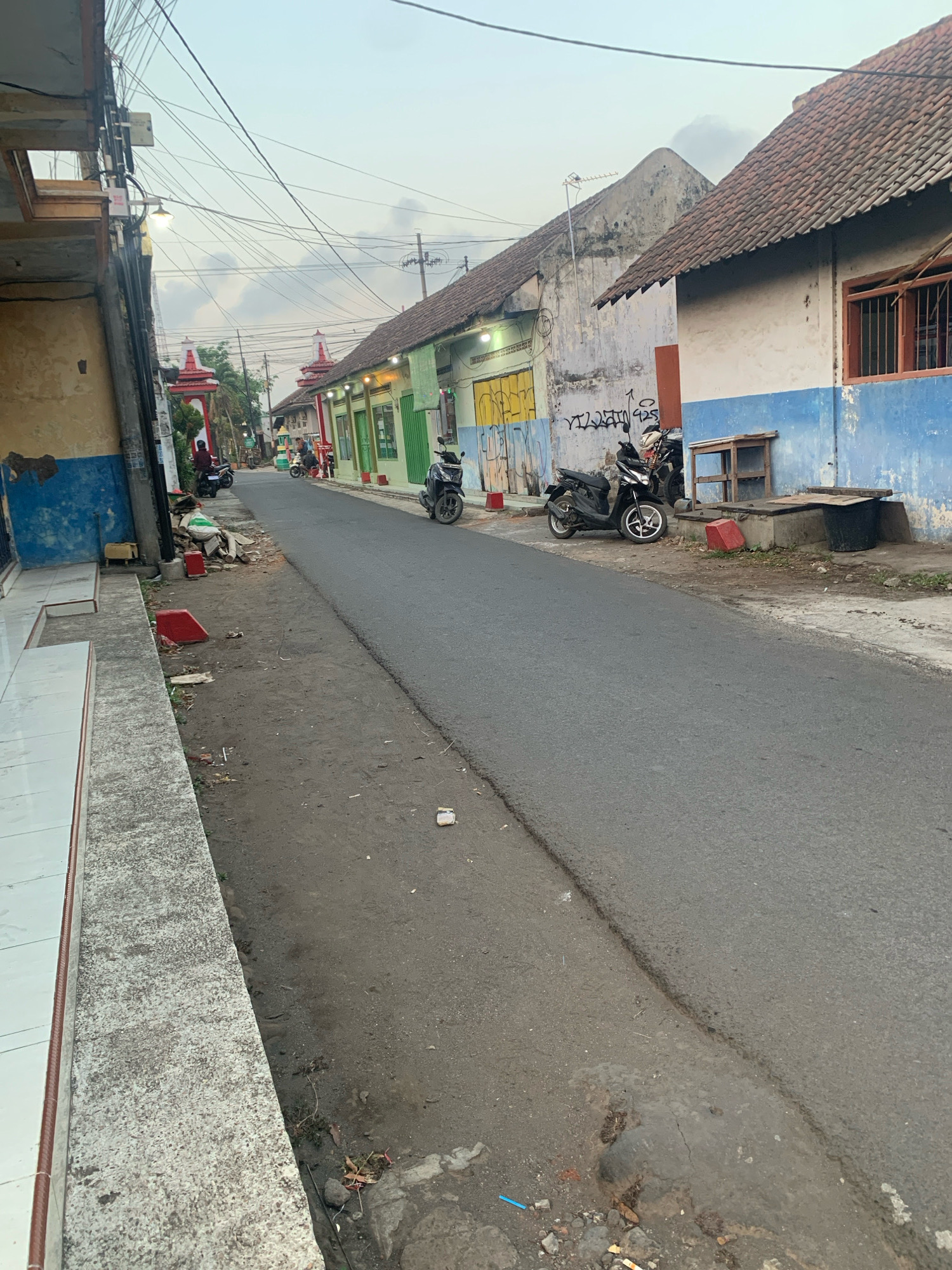 The image shows a street scene in Bululawang, Indonesia. The street is paved and appears to be in a residential or commercial area with buildings lining both sides. Several motorcycles are parked along the street. The buildings have varied facades, with some showing signs of age and others appearing more recently painted. One building on the right has a blue and white lower section and a tiled roof. There are overhead power lines and a street lamp visible. The ground along the side of the street is a mix of dirt and some debris. The sky is overcast, suggesting either early morning or late afternoon. There are a few people visible in the distance, seemingly engaged in everyday activities. The overall impression is of a functional street within a settled community in Bululawang.